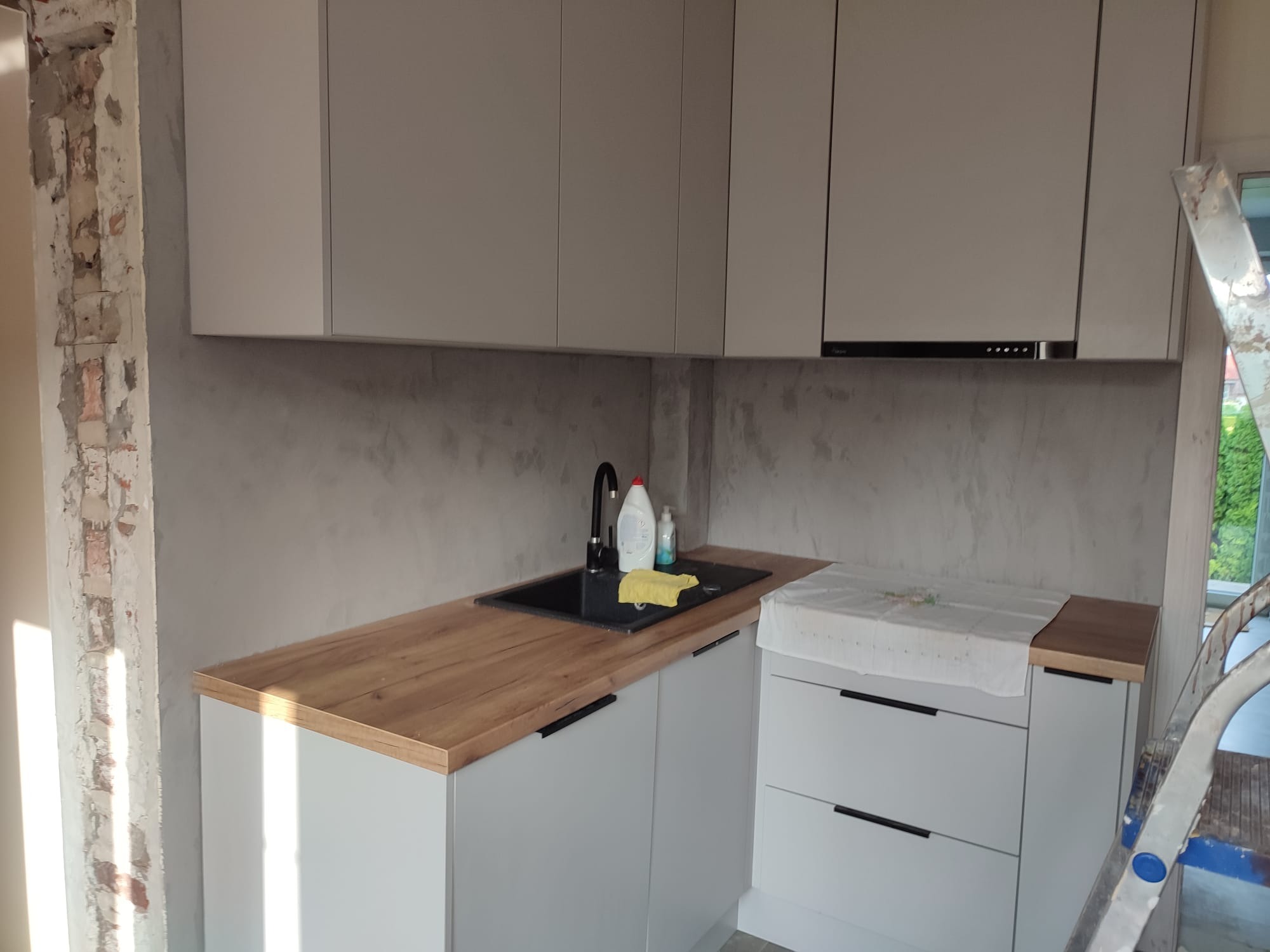 Jasna kuchnia z drewnianym blatem i czarnym zlewem, fragment ściany z odsłoniętą cegłą, drabina w rogu. Widok na etap wykończenia wnętrza.