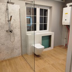 ER-GLAZ Rafał Kozłowski - Nowoczesna łazienka z kabiną typu walk-in, beżowymi płytkami podłogowymi imitującymi drewno i wiszącą toaletą. Widoczny bojler i okno.