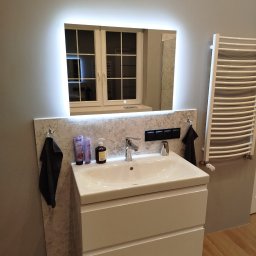 ER-GLAZ Rafał Kozłowski - Nowoczesna łazienka: biała szafka z umywalką, podświetlane lustro z odbiciem okna, grzejnik drabinkowy i akcesoria łazienkowe na tle szarej ściany i drewnianej podłogi.