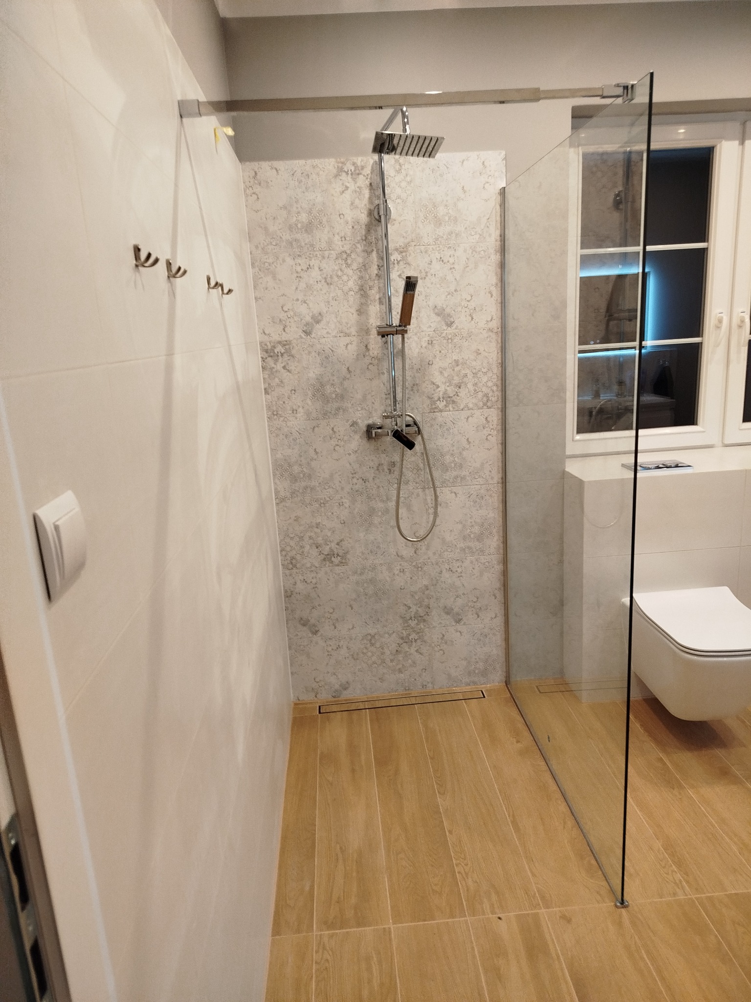 Nowoczesna kabina prysznicowa walk-in z deszczownicą i szklaną ścianką, wykończona płytkami w odcieniach bieli i beżu, z drewnopodobną podłogą i widoczną toaletą.