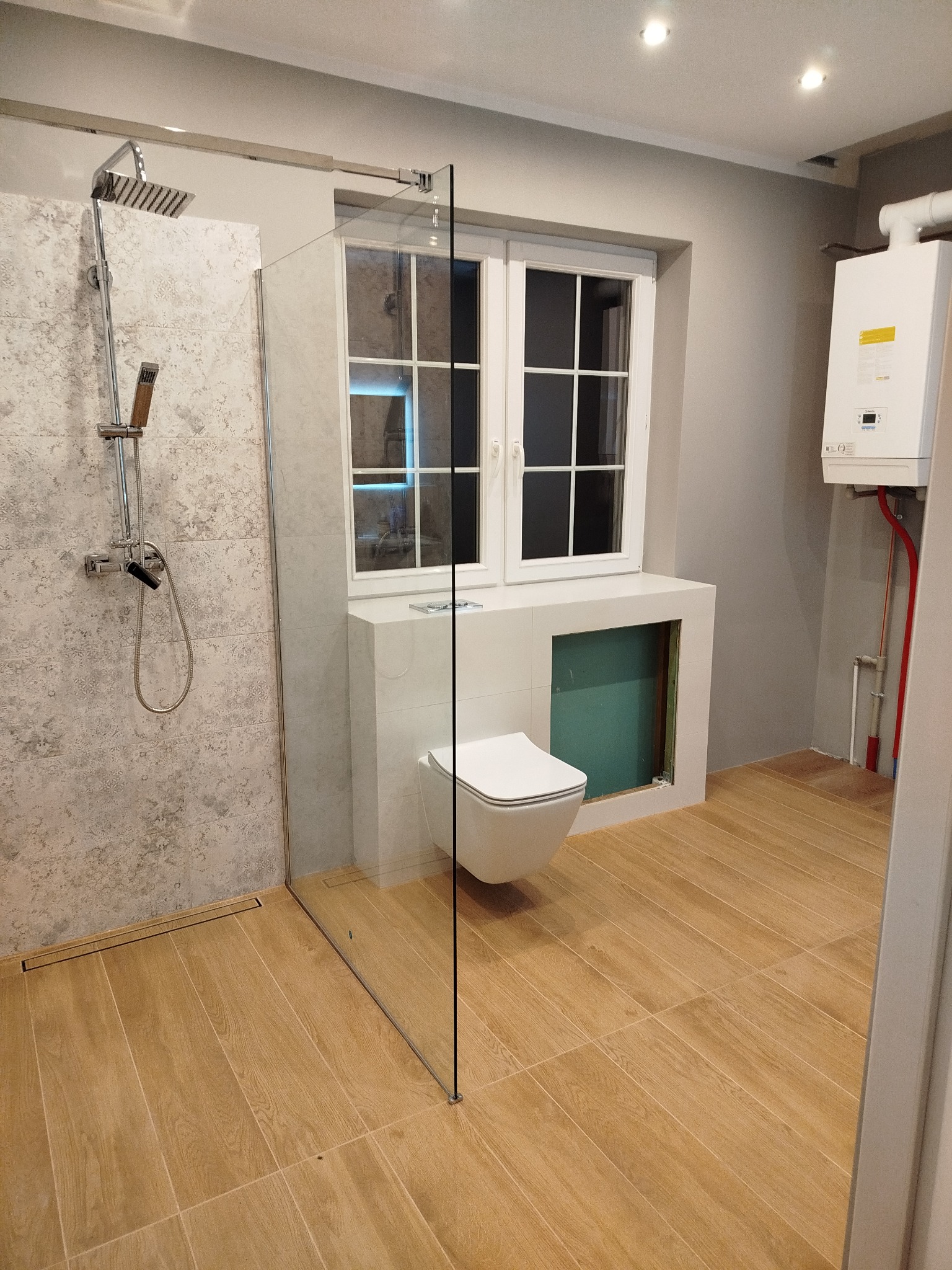 Nowoczesna łazienka z kabiną typu walk-in, beżowymi płytkami podłogowymi imitującymi drewno i wiszącą toaletą. Widoczny bojler i okno.