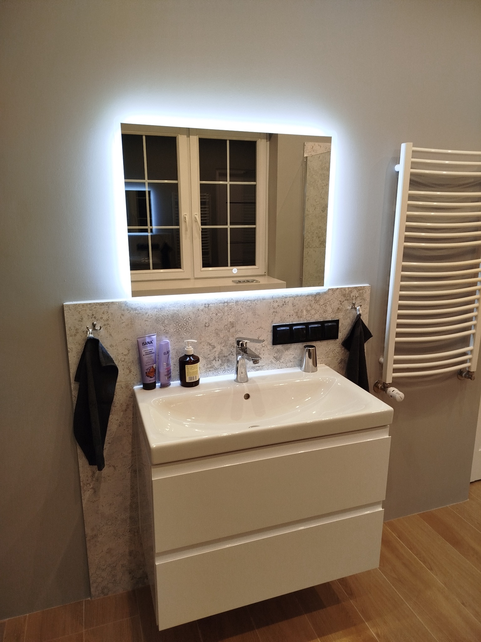 Nowoczesna łazienka: biała szafka z umywalką, podświetlane lustro z odbiciem okna, grzejnik drabinkowy i akcesoria łazienkowe na tle szarej ściany i drewnianej podłogi.