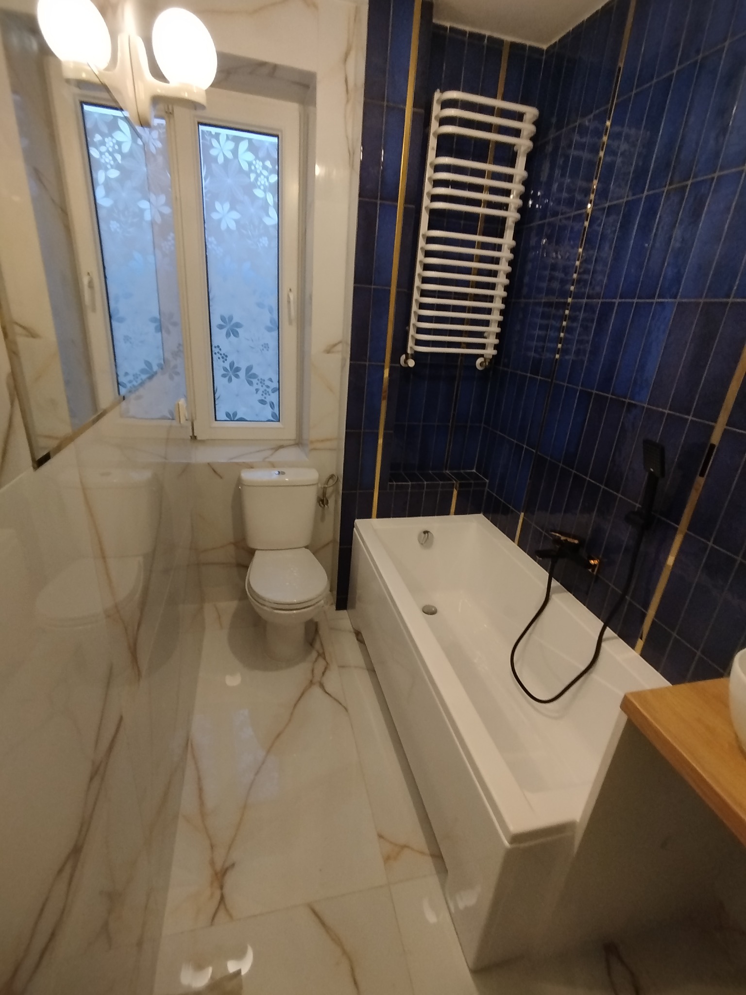 Wykończona łazienka z białą armaturą, niebieskimi płytkami z złotymi akcentami i grzejnikiem drabinkowym. Widok na okno z dekoracyjną folią.