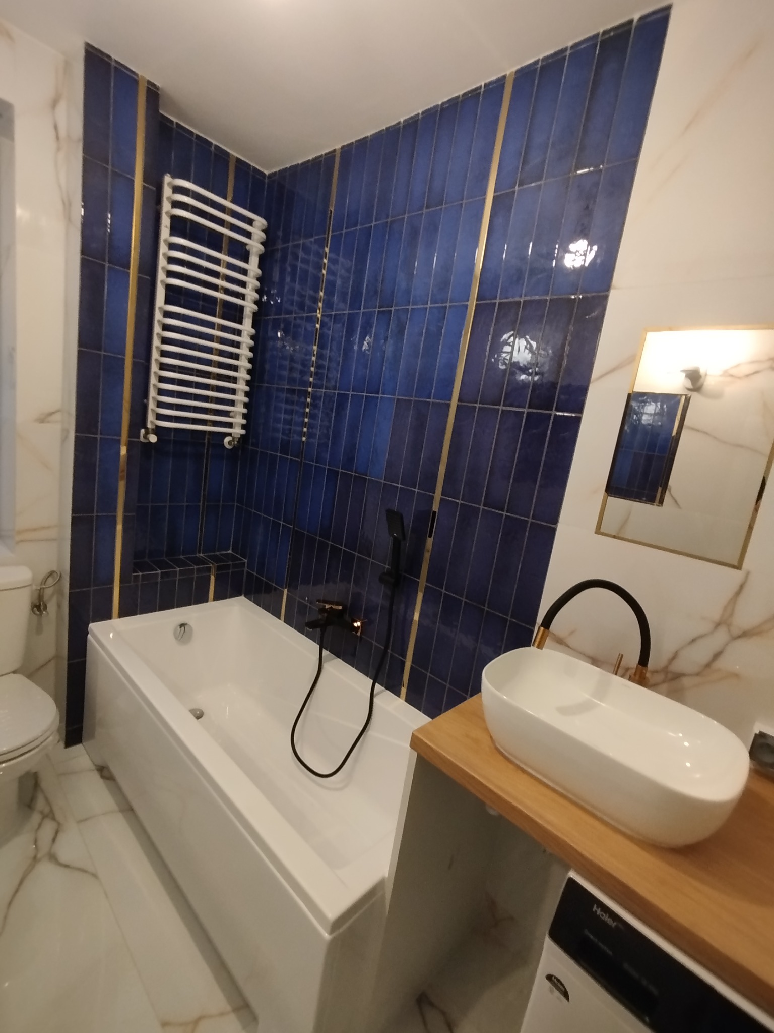 Nowoczesna łazienka z wanną, umywalką nablatową i grzejnikiem drabinkowym na tle kobaltowych płytek z złotymi akcentami. Marmurowe płytki podłogowe.
