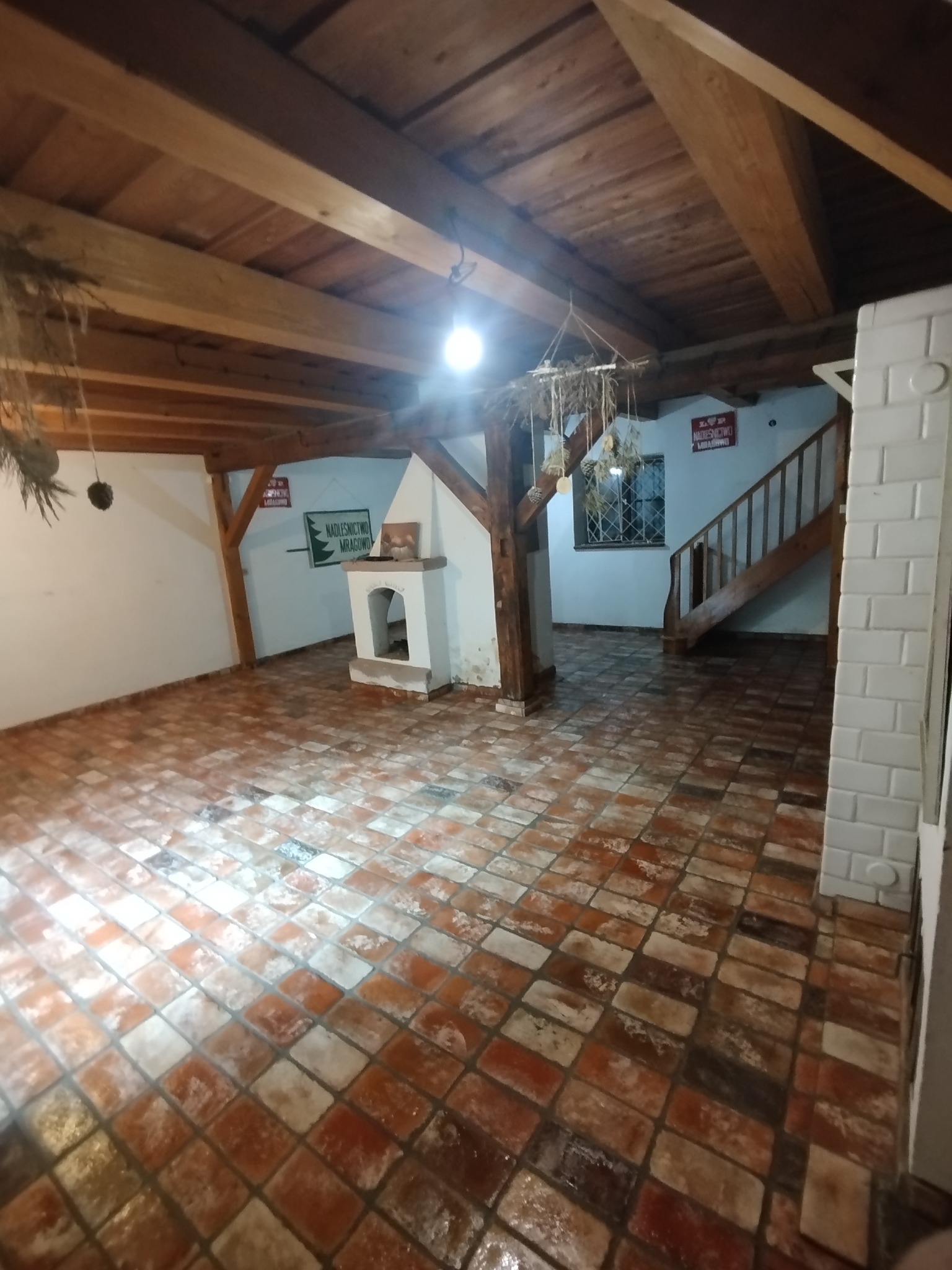 Rustykalne wnętrze z drewnianym stropem, ceglaną podłogą i kominkiem. Widoczne schody i dekoracje z suszonych roślin. Charakterystyczny styl wykończenia.