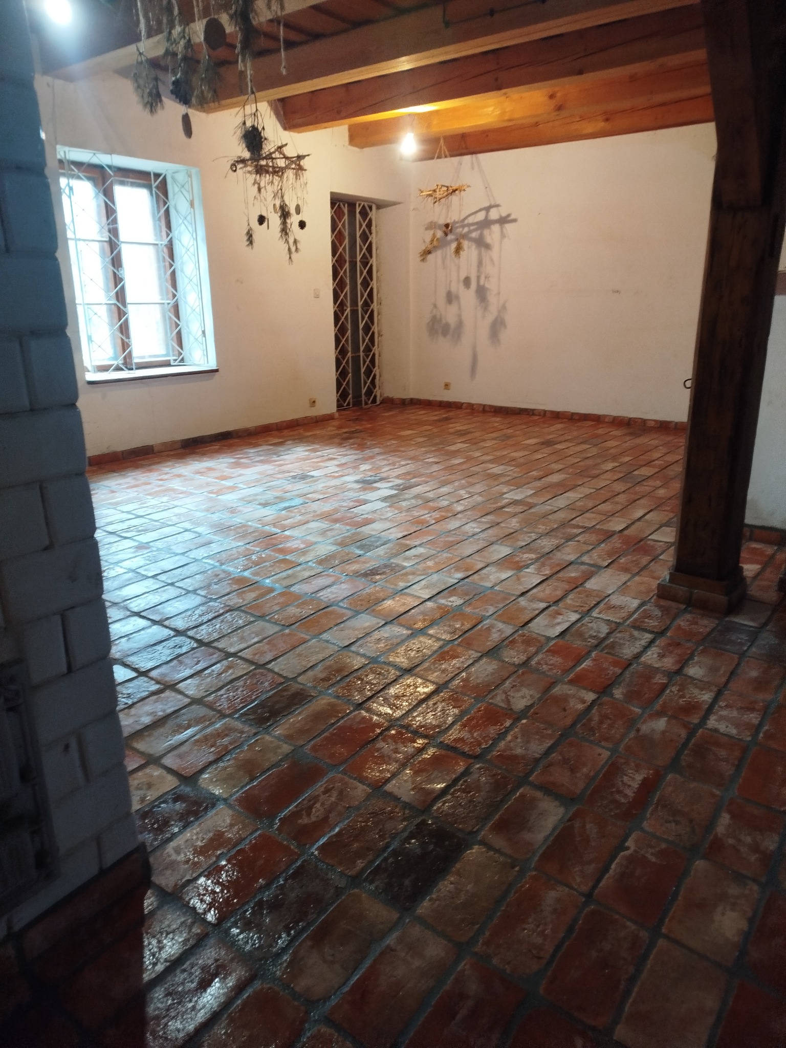 Rustykalne, czerwono-brązowe płytki podłogowe ułożone w kwadraty, w przestronnym wnętrzu z drewnianymi belkami stropowymi i kolumną. Widoczne dekoracje wiszące.
