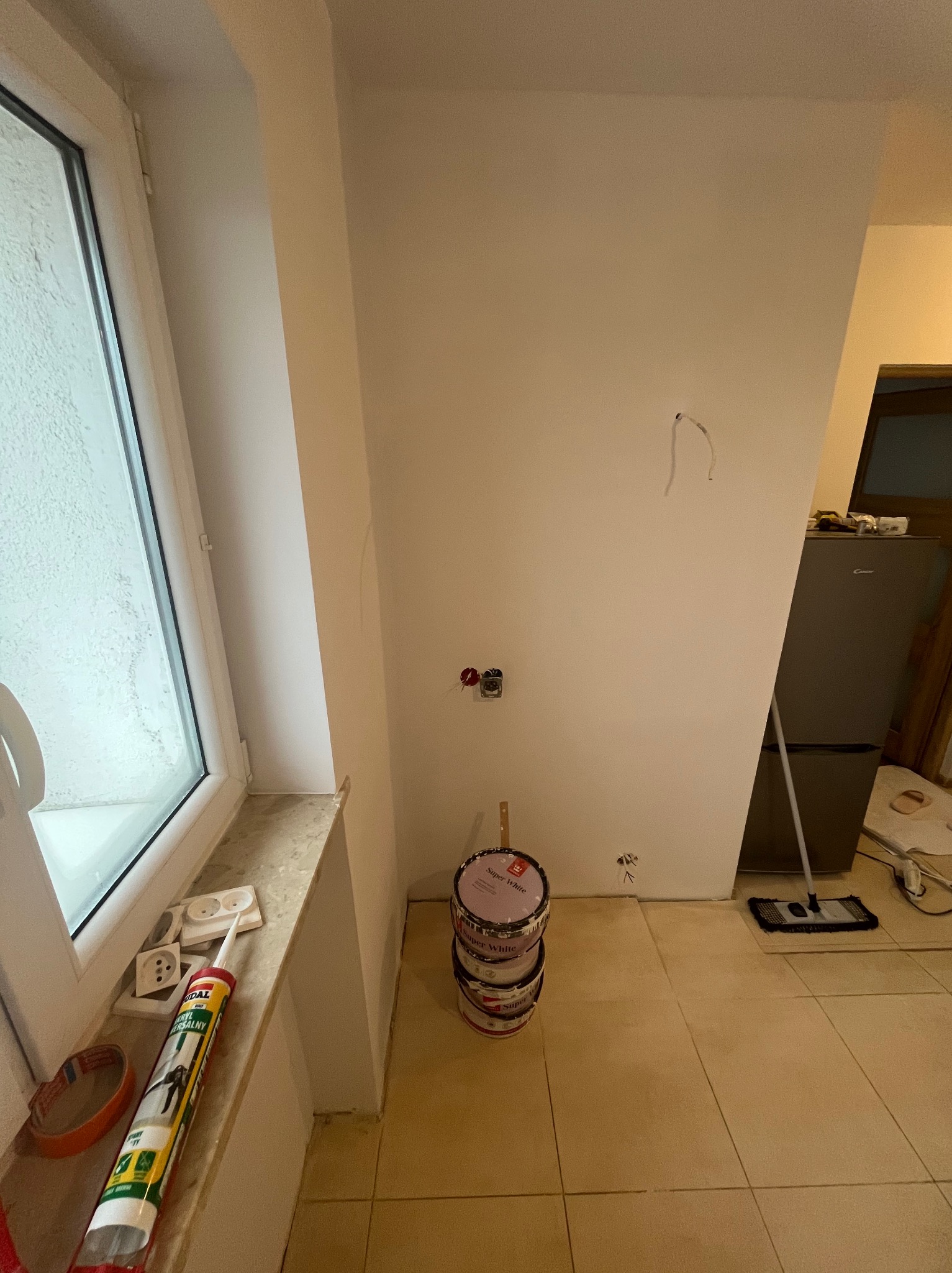 Biała ściana w trakcie malowania, widoczne puszki z farbą Super White, narzędzia malarskie i gniazdka elektryczne. Przy oknie leży silikon i taśma malarska.