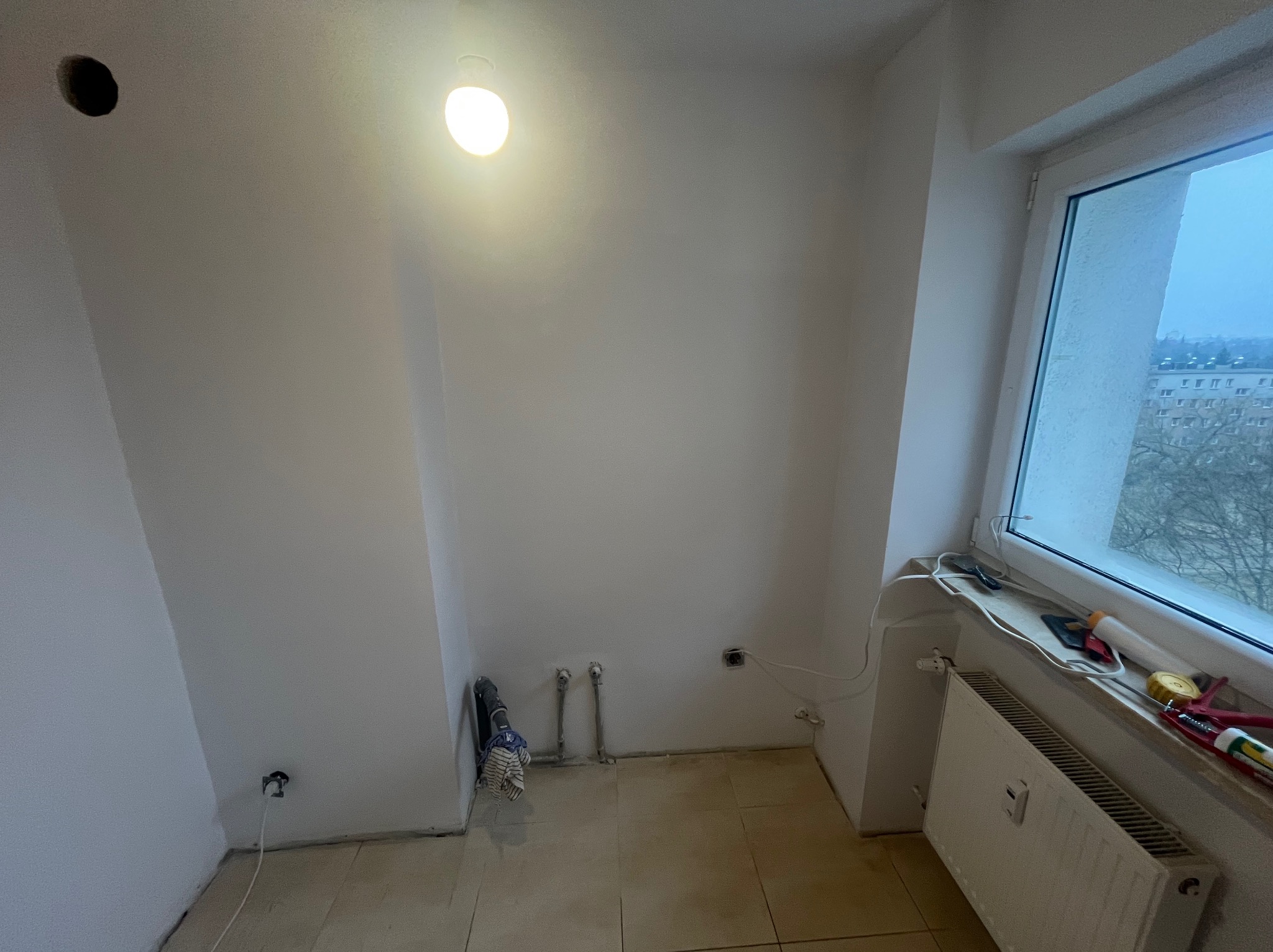 Wnętrze pokoju po szpachlowaniu ścian, z widocznymi instalacjami grzewczymi i elektrycznymi. Na parapecie narzędzia. Widok na zewnątrz przez okno.