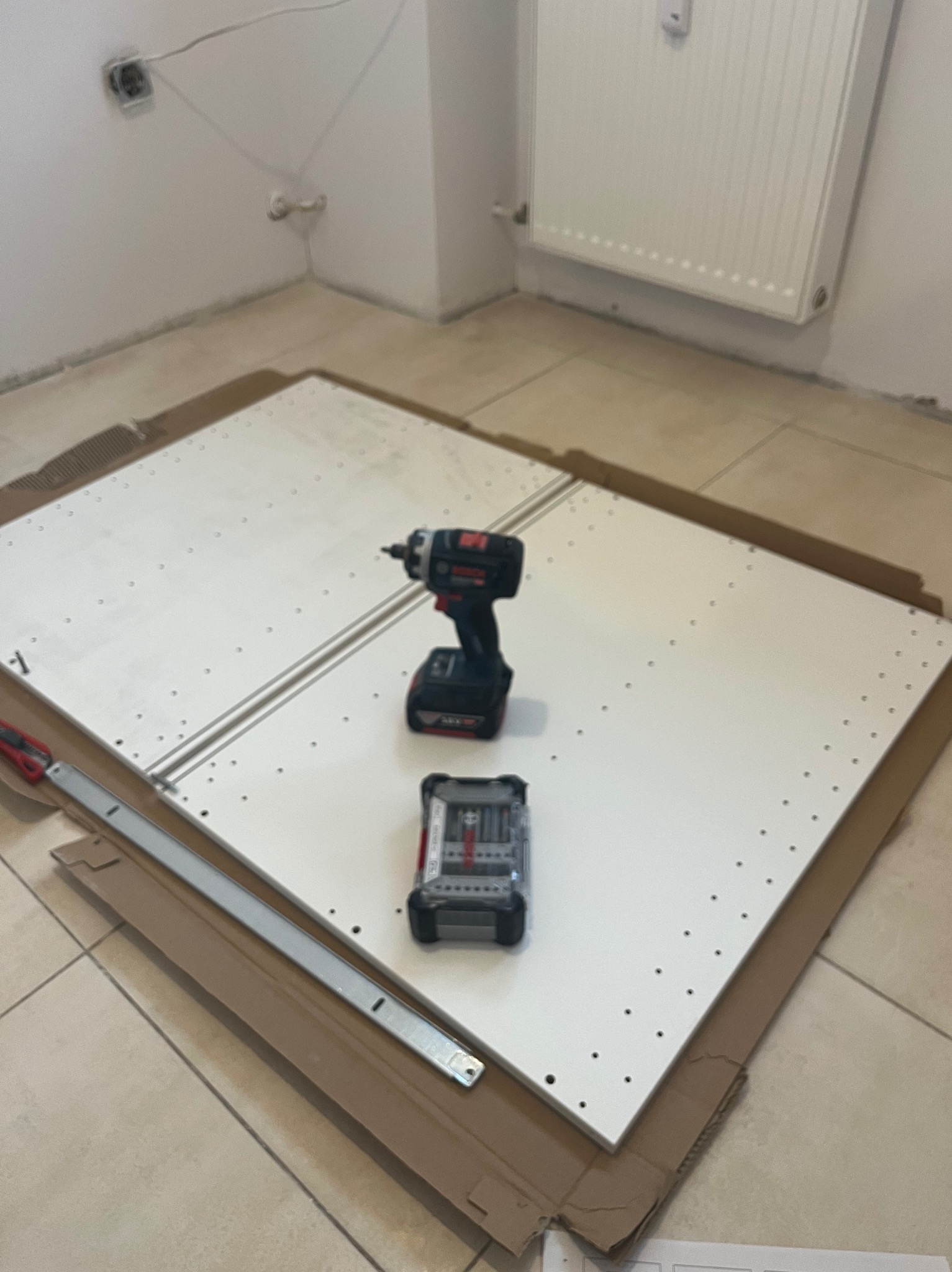 Biała płyta meblowa z nawierconymi otworami, wkrętarka akumulatorowa i zestaw bitów na podłodze w trakcie montażu. Widoczne elementy montażowe i narzędzia.
