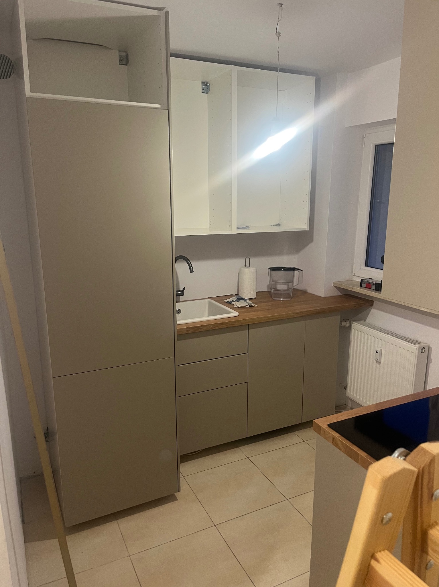 Montaż nowoczesnej, beżowej zabudowy kuchennej z białymi szafkami wiszącymi, widoczny zlew, bateria i fragment okna. Na podłodze płytki. Etap prac wykończeniowych.