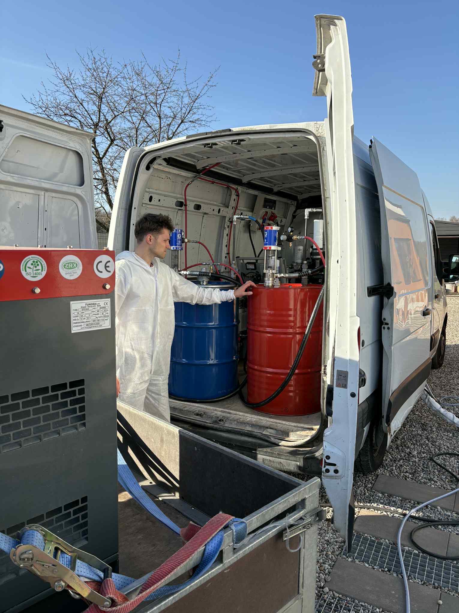 Pracownik w kombinezonie przy otwartym furgonie z beczkami i sprzętem do natrysku pianki PUR. Widoczne detale urządzenia i otoczenie placu budowy.