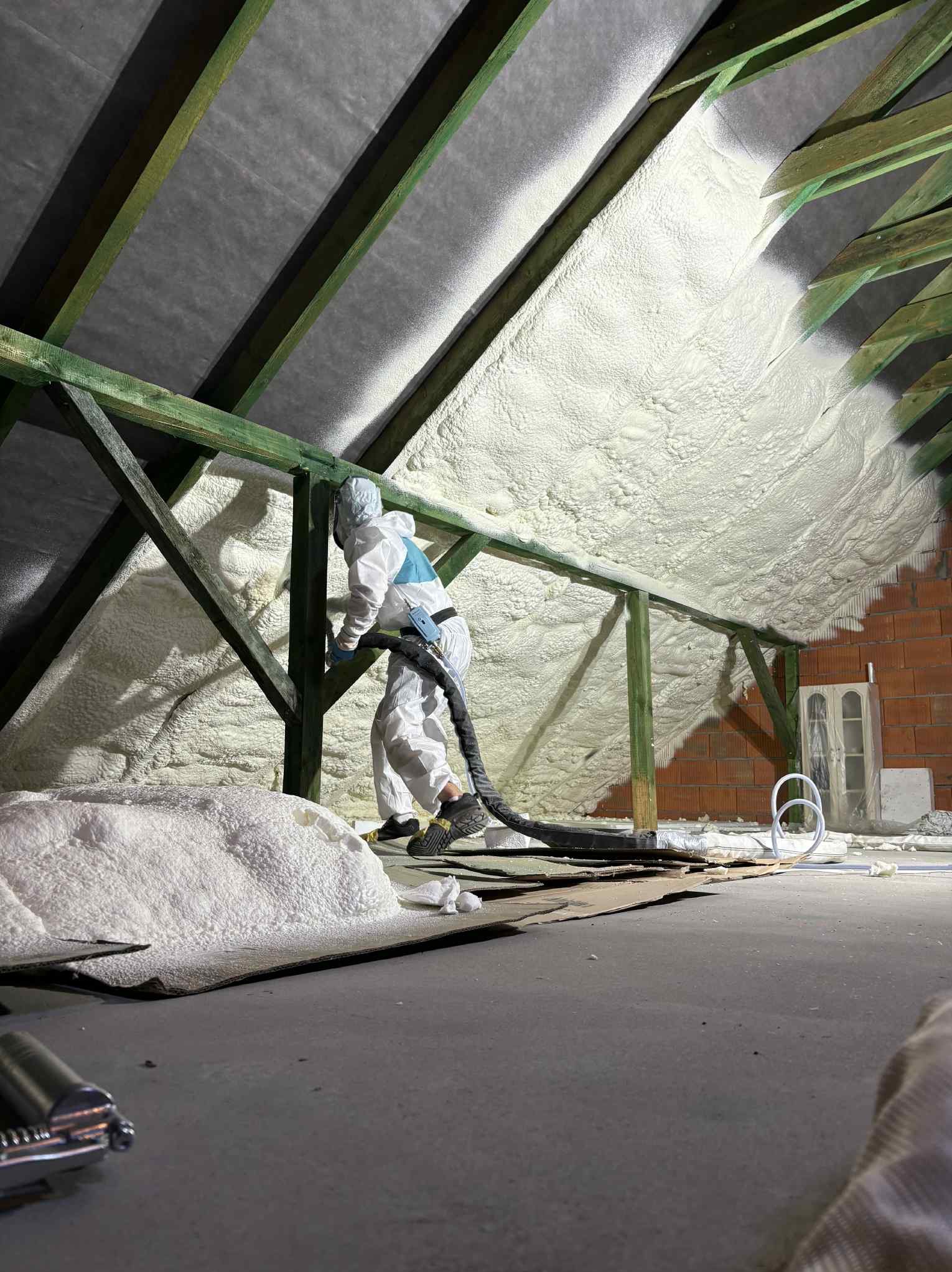 Pracownik w kombinezonie aplikuje pianę PUR na poddaszu, widoczne drewniane elementy konstrukcyjne i fragment ściany z cegły. Izolacja natryskowa w trakcie realizacji.