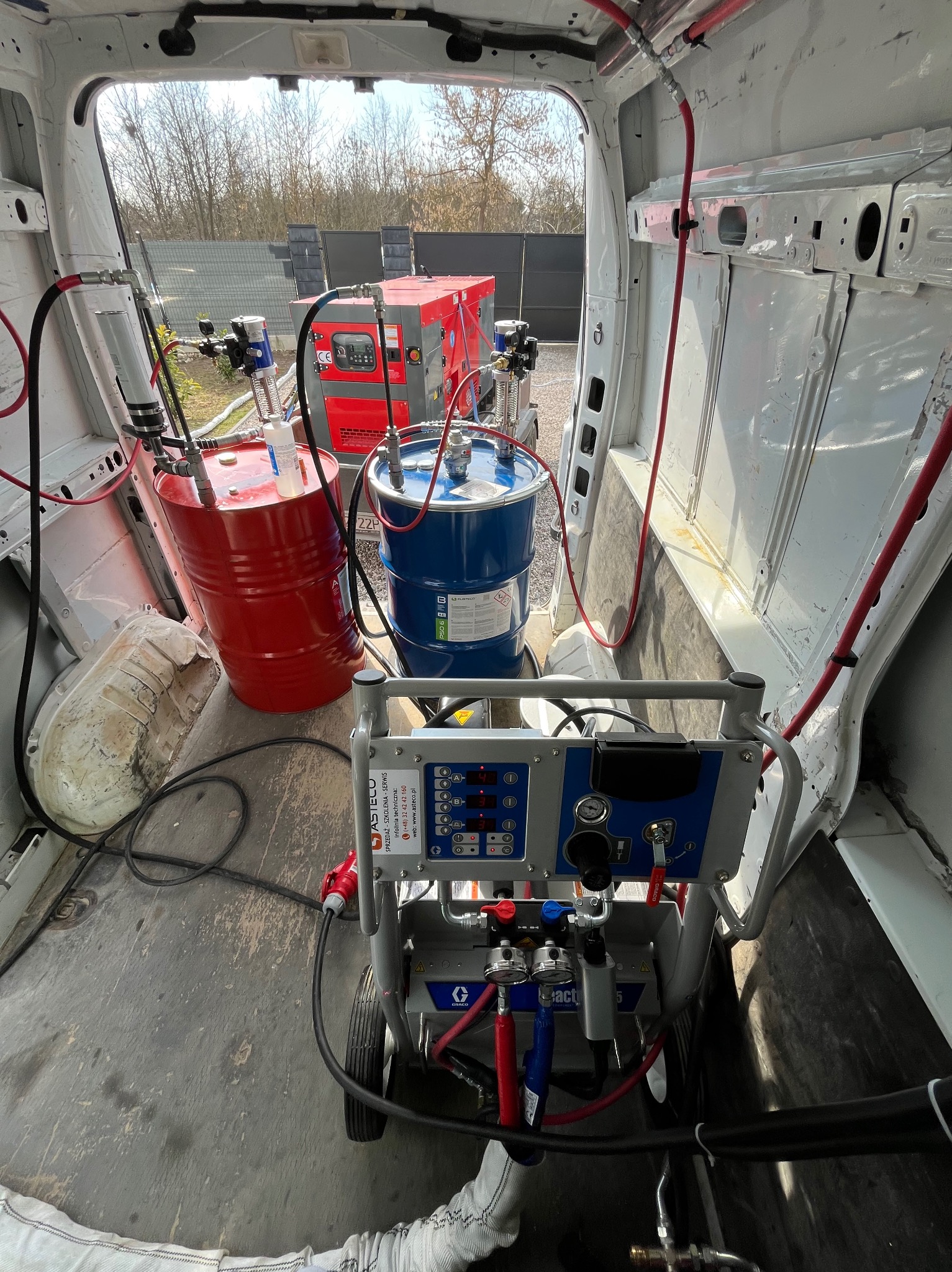 Wnętrze furgonetki z maszyną do natrysku pianki PUR, zbiornikami z komponentami i agregatem prądotwórczym. Widoczne przewody i elementy sterujące.