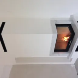 Nowoczesna zabudowa z białych płyt i marmurowymi akcentami, z widocznym płomieniem wewnątrz paleniska, oraz czarnym, geometrycznym wlotem powietrza po lewej stronie.