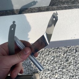 WINEXO - Ręka trzyma metalowe elementy okienne, prawdopodobnie podczas naprawy lub montażu. Widoczne detale mocowań i profil okienny na tle szarego żwiru.