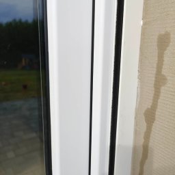 WINEXO - Szczegółowe ujęcie uszczelki okiennej w białej ramie, z widoczną fakturą ściany i fragmentem szyby z odbiciem zieleni na zewnątrz.