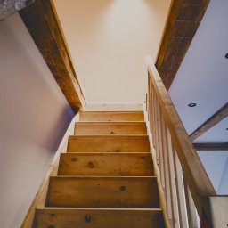 Djl.Gk.Pro Daniel Larek - Drewniane schody z balustradą, widok od dołu, prowadzące na górę. Widoczne drewniane belki stropowe i oświetlenie sufitowe. Naturalne wykończenie drewna.