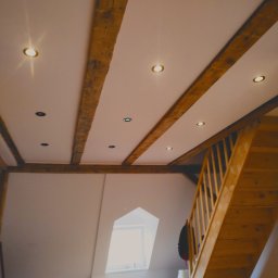 Djl.Gk.Pro Daniel Larek - Jasne wnętrze poddasza z drewnianymi belkami stropowymi i schodami. Sufit z wpuszczonymi punktowymi lampami. Okno dachowe w ścianie. Widok z dołu.