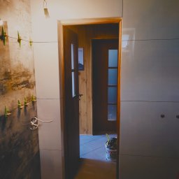 Djl.Gk.Pro Daniel Larek - Ściana łazienki w trakcie wykańczania: połowa ściany z surowym betonem, druga połowa z błyszczącymi, szarymi płytkami. Widoczne krzyżyki glazurnicze i wiadro z narzędziami.