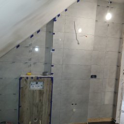 Djl.Gk.Pro Daniel Larek - Ściana w łazience w trakcie układania szarych płytek, zabezpieczonych niebieską taśmą. Widoczna zabudowa stelaża WC z drewnopodobną okleiną i wystające kable.