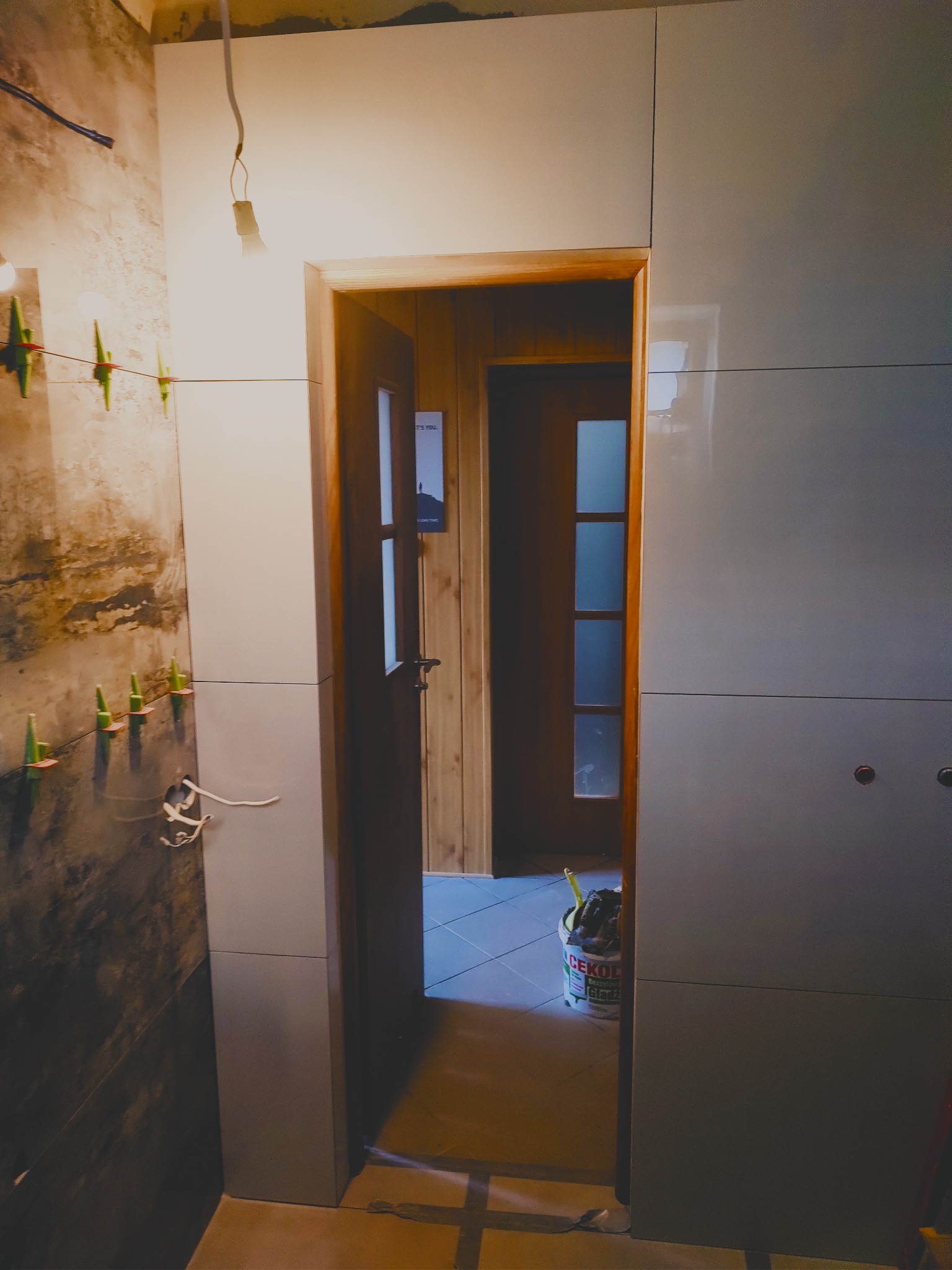 Ściana łazienki w trakcie wykańczania: połowa ściany z surowym betonem, druga połowa z błyszczącymi, szarymi płytkami. Widoczne krzyżyki glazurnicze i wiadro z narzędziami.