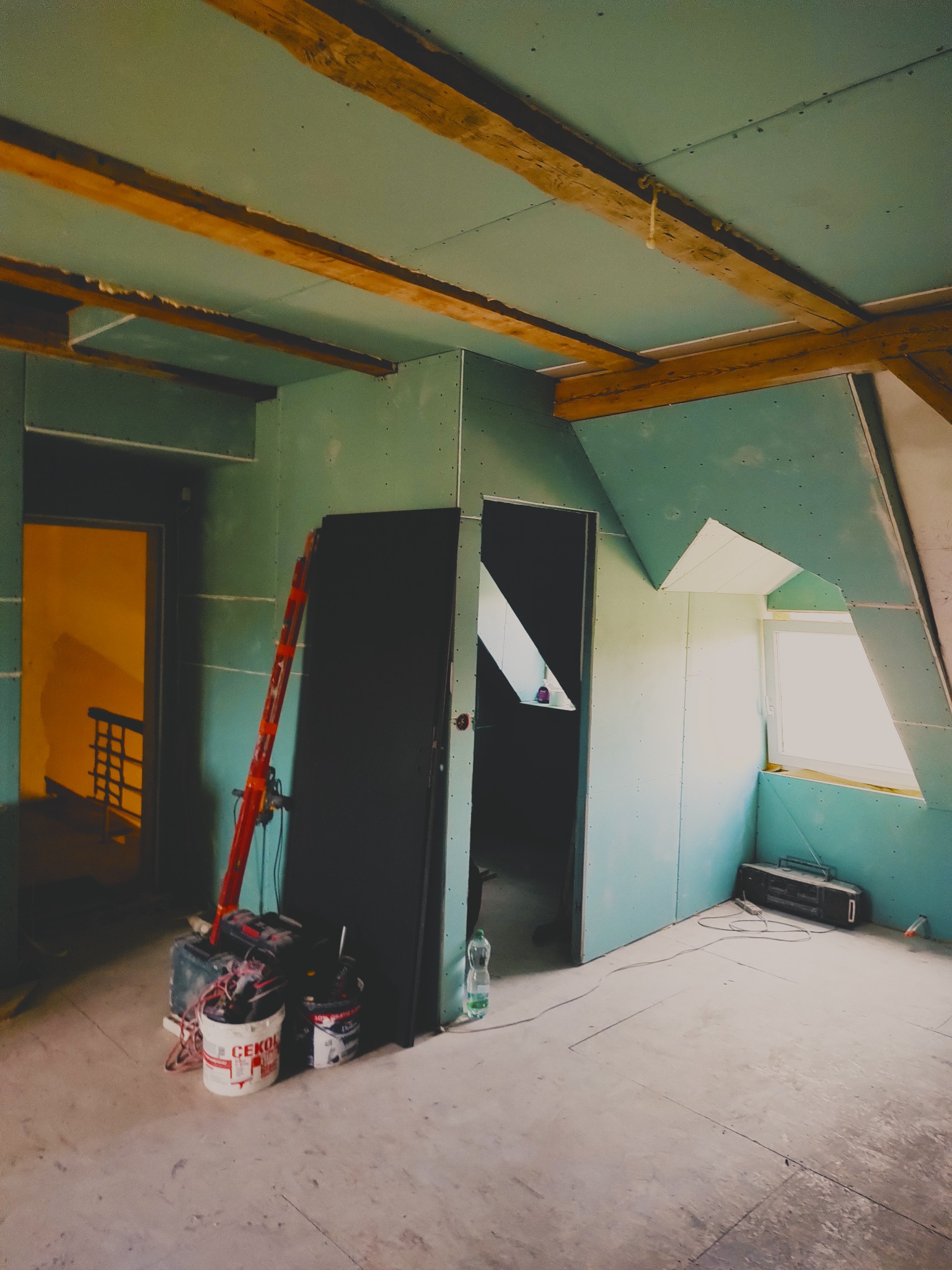 Poddasze w trakcie adaptacji: ściany obłożone płytami gipsowo-kartonowymi, widoczne drewniane belki stropowe, drabina i narzędzia malarskie. Otwór okienny w skosie dachu.