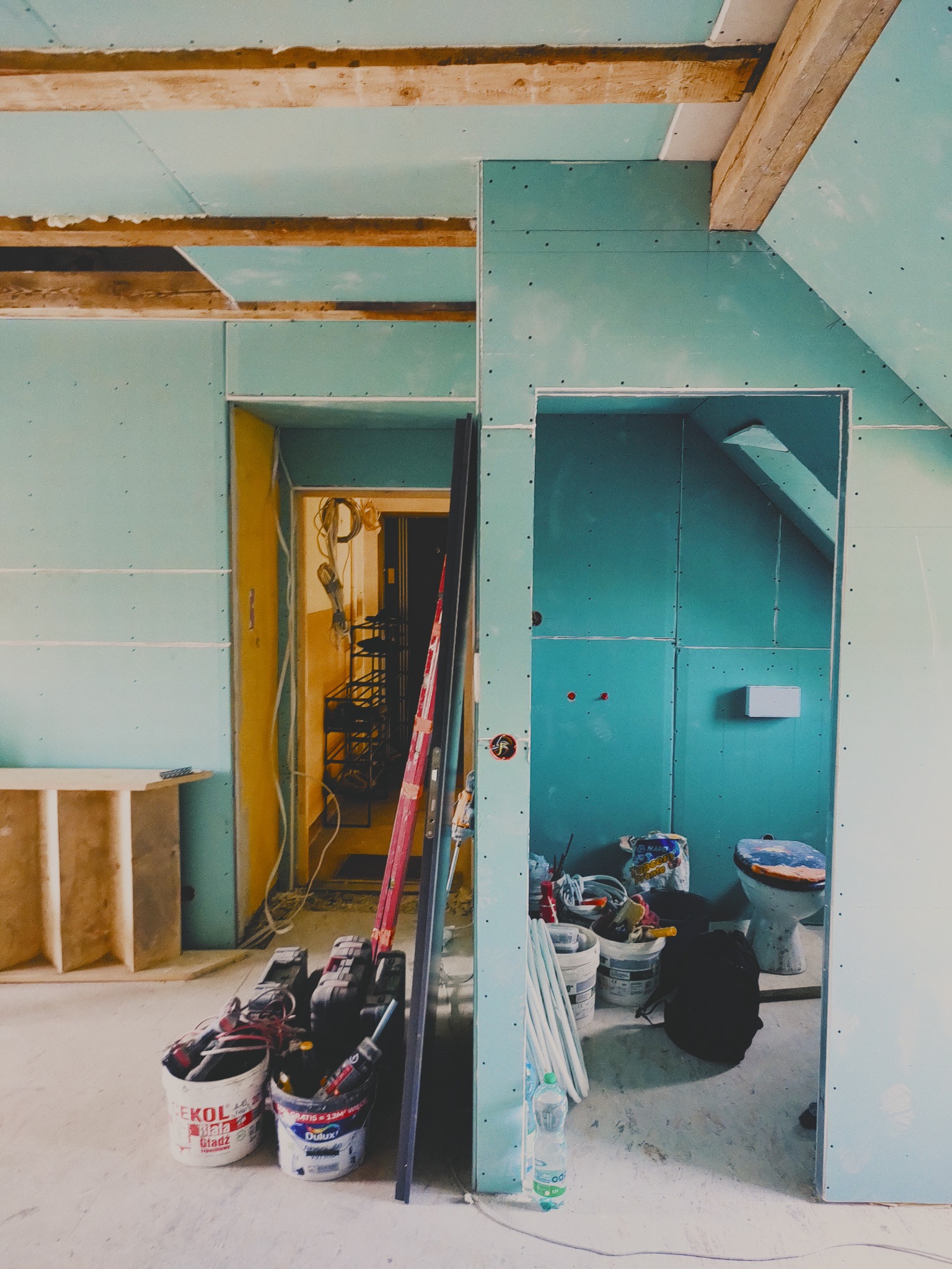 Remont poddasza: ściany z płyt karton-gips, widoczne narzędzia i materiały budowlane, przygotowane pomieszczenie łazienki z toaletą. Prace wykończeniowe w toku.