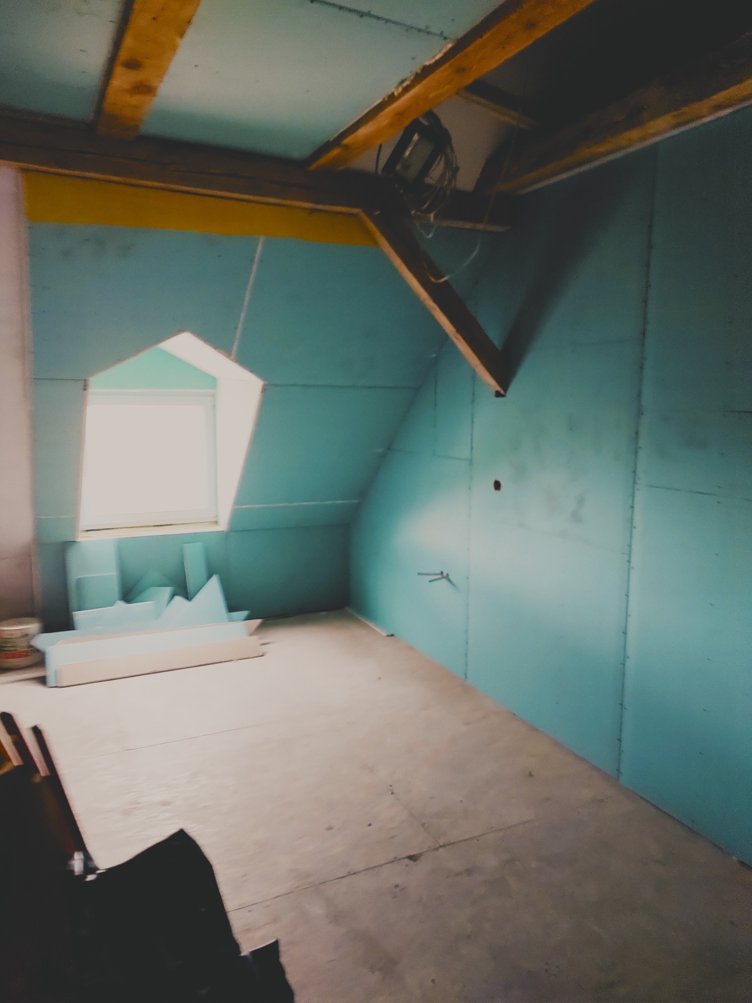 Poddasze z oknem dachowym w trakcie prac wykończeniowych. Ściany obłożone płytami kartonowo-gipsowymi w kolorze turkusowym, widoczne drewniane elementy konstrukcyjne.