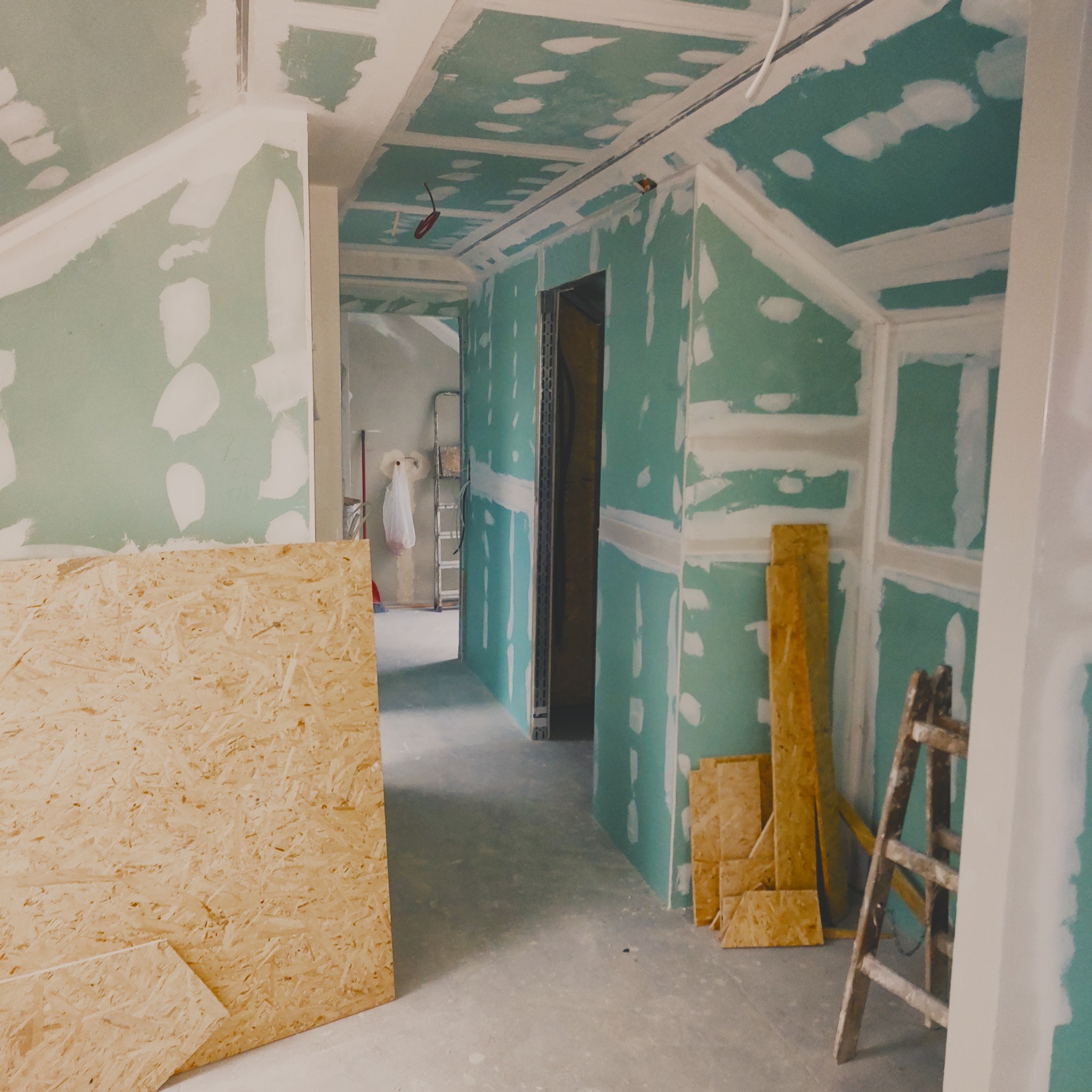 Wnętrze w trakcie prac: ściany i sufit pokryte płytami kartonowo-gipsowymi, widoczne łączenia i szpachlowanie. Na podłodze płyta OSB i drabina malarska.