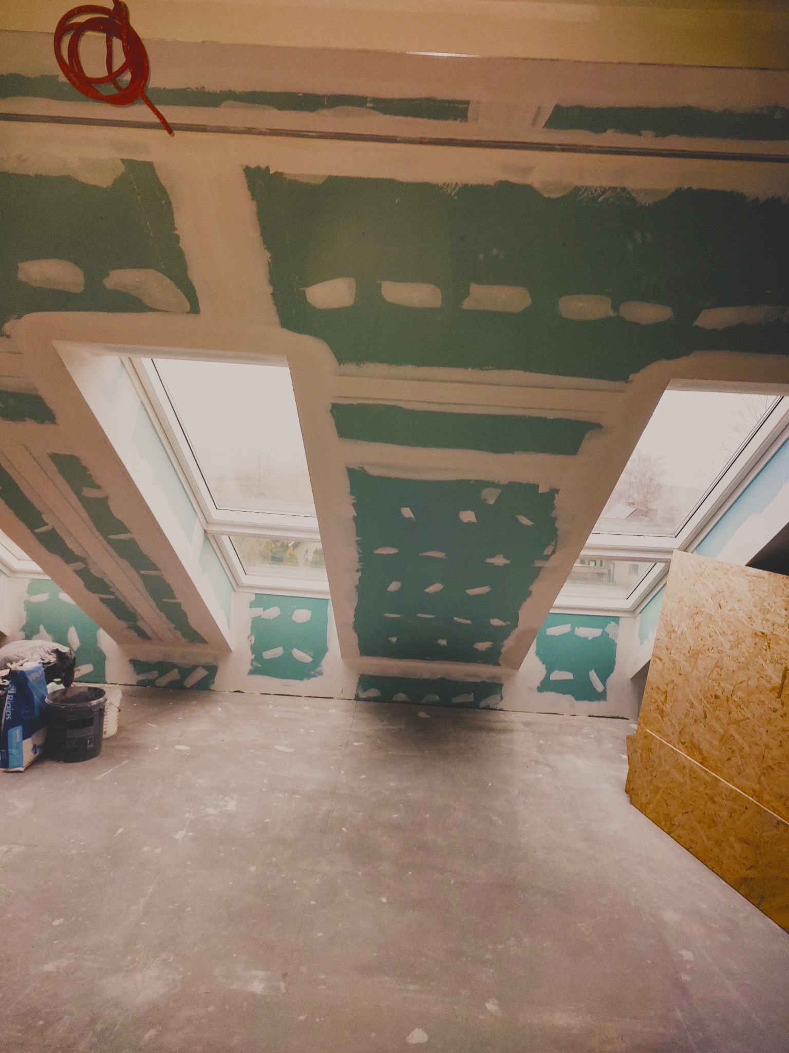 Poddasze z oknami dachowymi w trakcie prac wykończeniowych. Widoczne płyty gipsowo-kartonowe i szpachlowane łączenia. Na podłodze widoczne materiały budowlane.