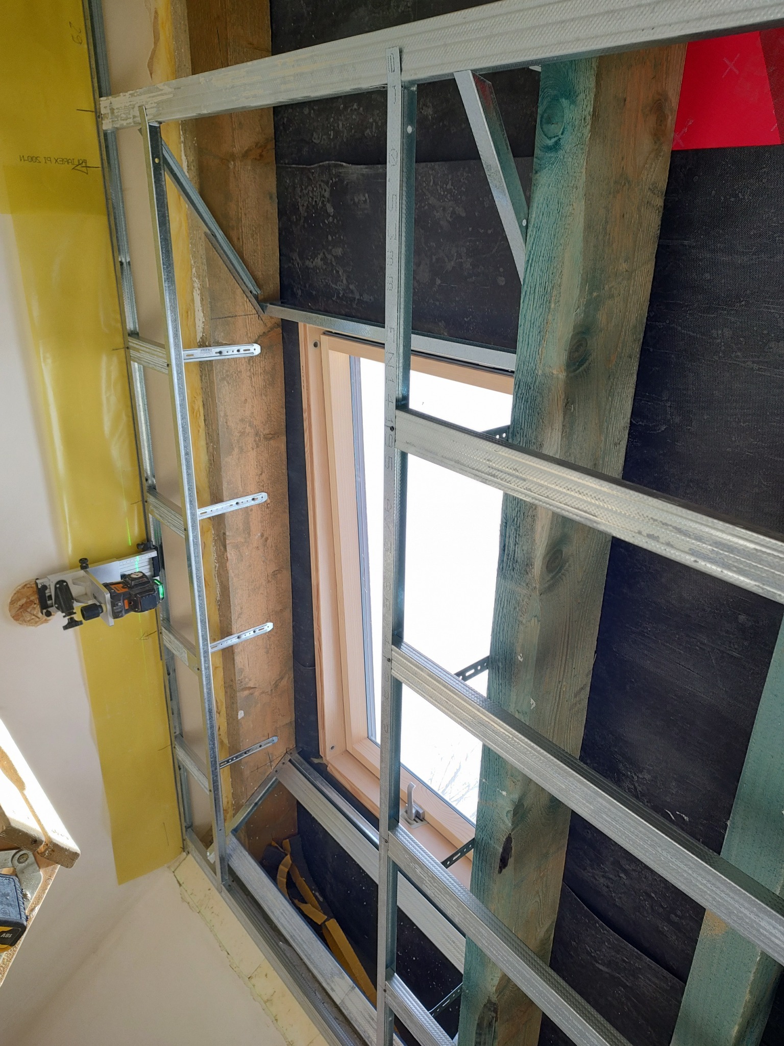 Montaż metalowej konstrukcji pod płyty gipsowo-kartonowe przy oknie dachowym, widoczny laser krzyżowy i drewniane elementy konstrukcyjne dachu.