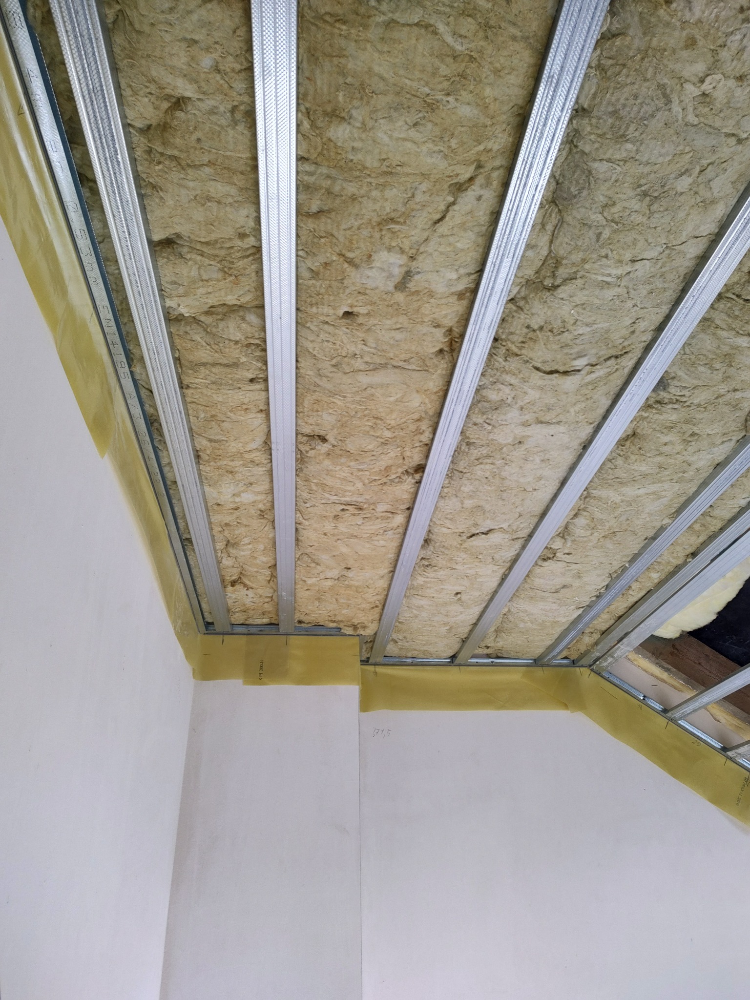 Montaż izolacji z wełny mineralnej w suficie podwieszanym, widoczne metalowe profile i folia paroizolacyjna na styku ściany i sufitu.