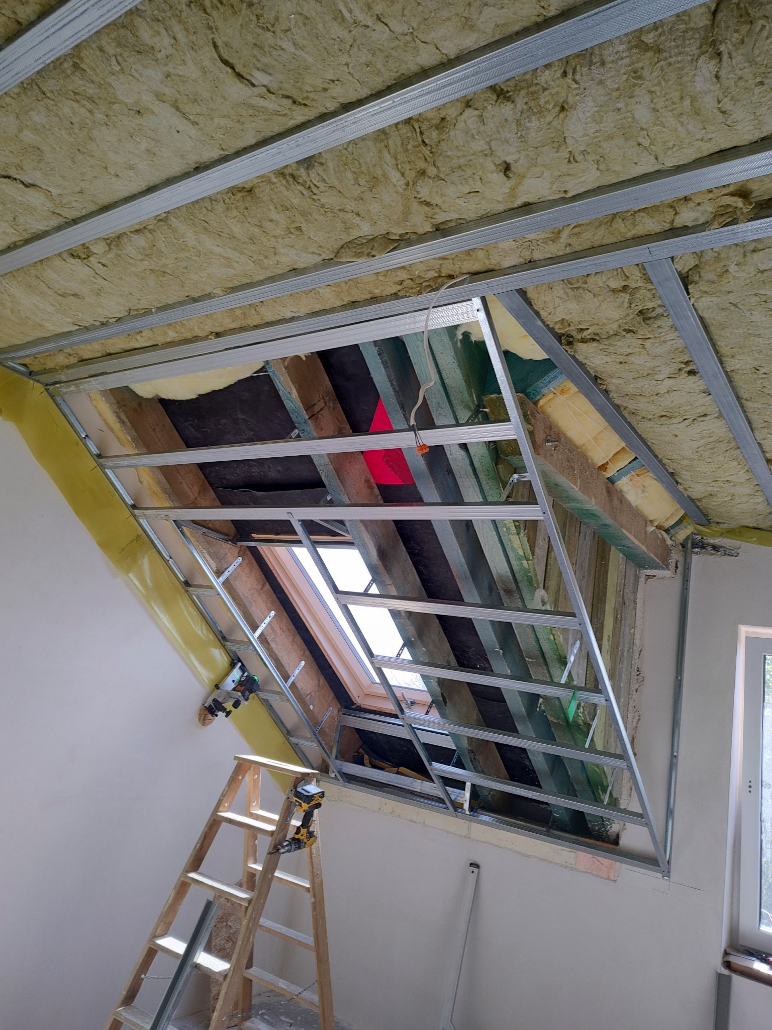 Skośny sufit z izolacją i metalową konstrukcją pod płyty, widok z dołu. Drabina oparta o ścianę, narzędzia na szczeblach. Okno dachowe w tle.