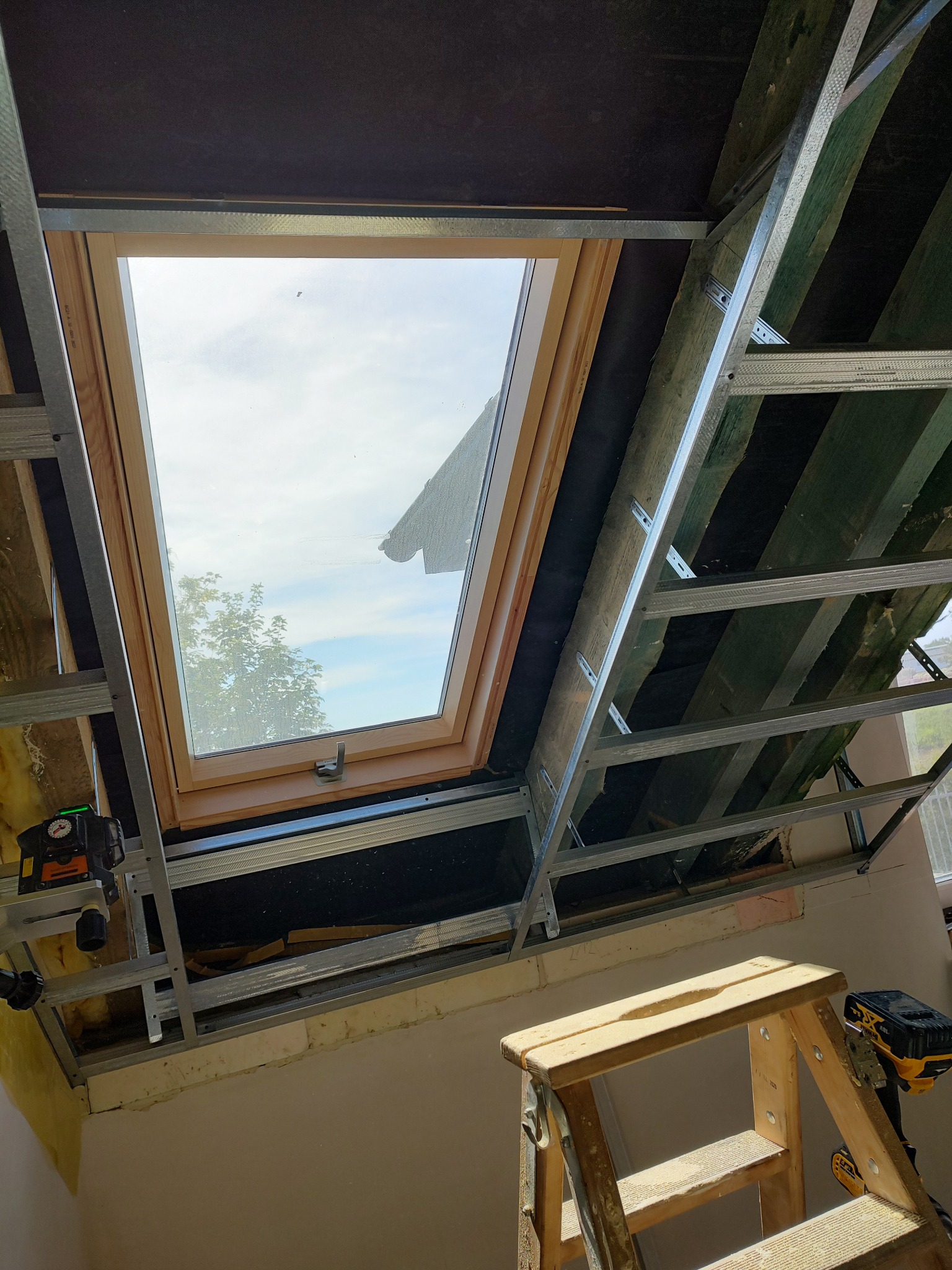 Montaż okna dachowego w konstrukcji z profili metalowych. Widoczne narzędzia i drabina. Prace wykończeniowe w trakcie realizacji.