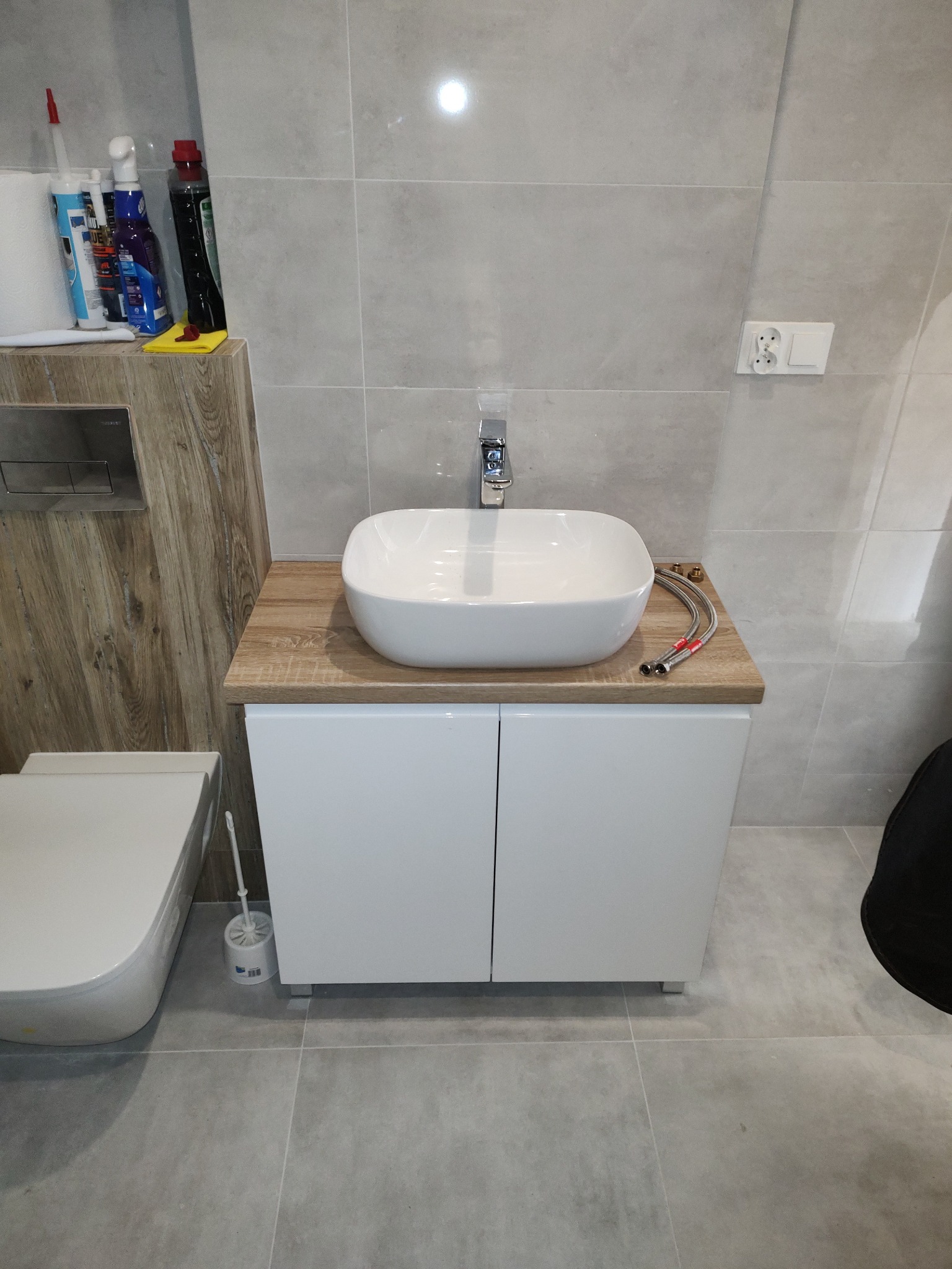 Biała umywalka nablatowa na szafce z blatem imitującym drewno, chromowana bateria, szare płytki na ścianie i podłodze. Widoczne wężyki do podłączenia wody.