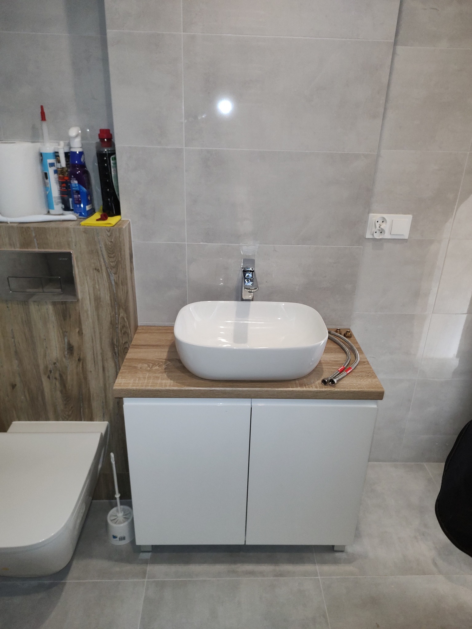 Nowoczesna łazienka z umywalką nablatową na szafce z drewnianym blatem, szare płytki na ścianach i podłodze. Widoczne wężyki do podłączenia baterii.