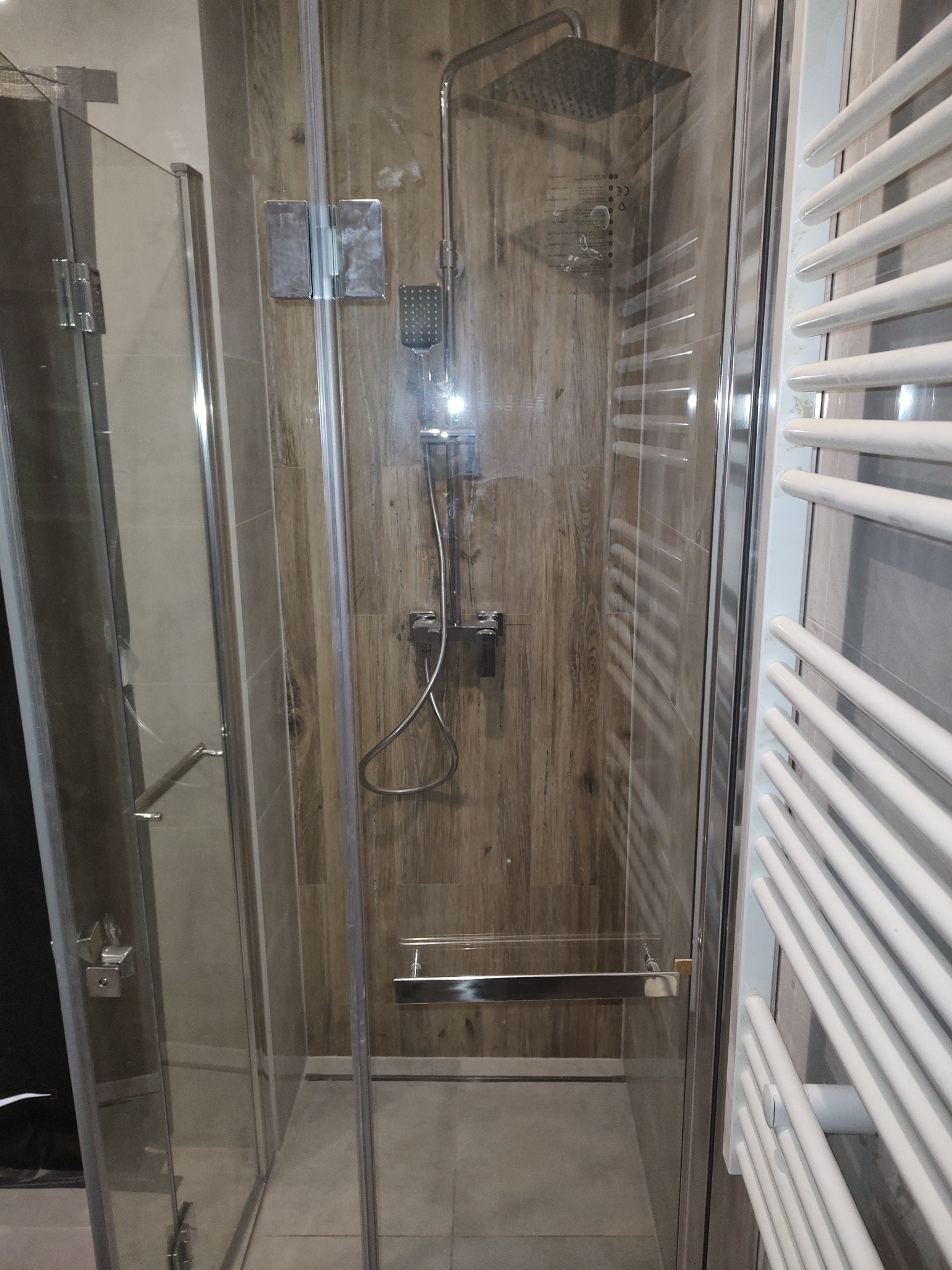 Kabina prysznicowa z deszczownicą i panelem imitującym drewno, widoczny grzejnik drabinkowy. Wykończenie łazienki w nowoczesnym stylu.