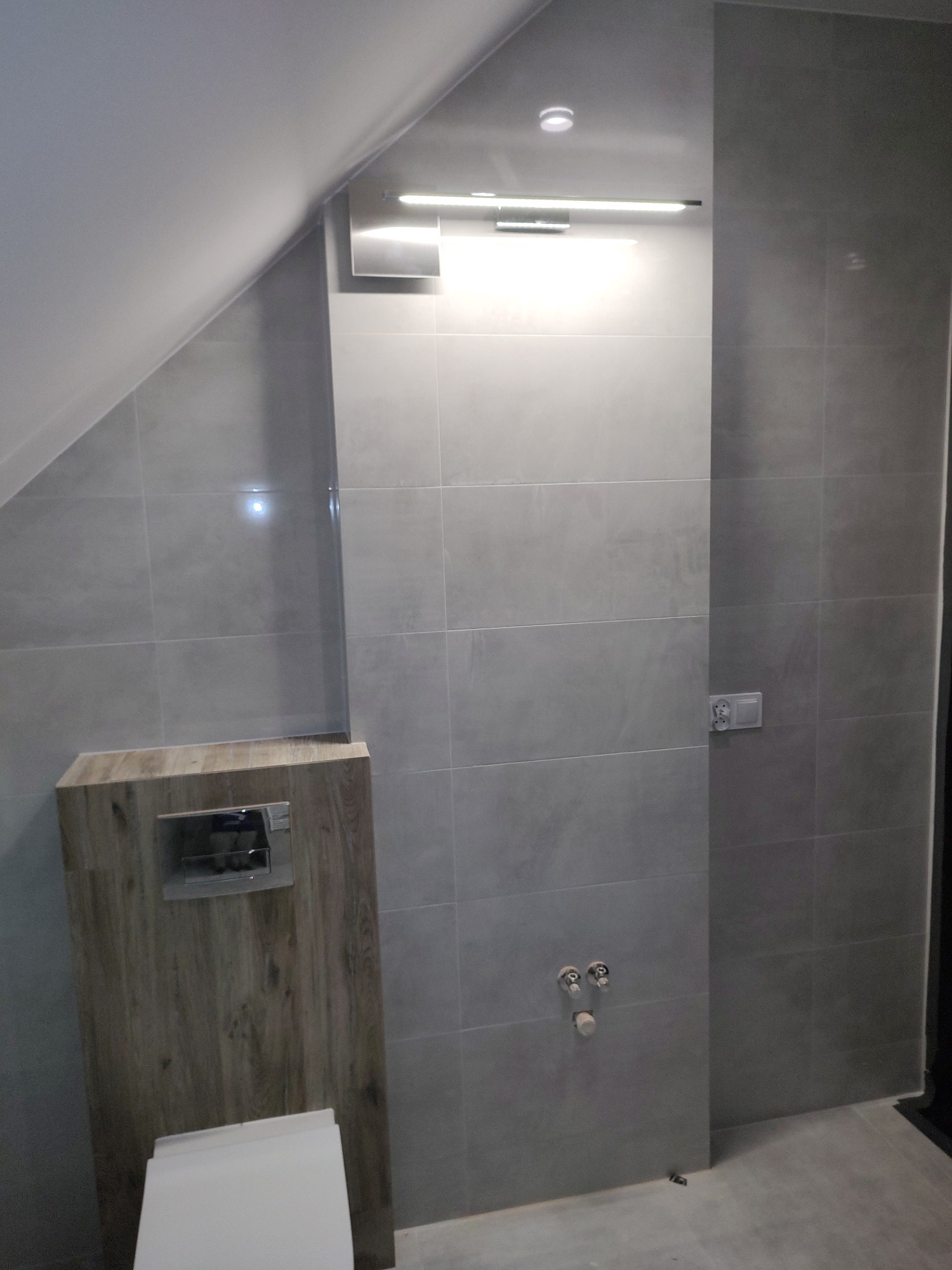 Wykończona łazienka na poddaszu: szare płytki, zabudowa WC z drewnopodobną okleiną, oświetlenie LED. Minimalistyczny design, widoczne przyłącza wodne.