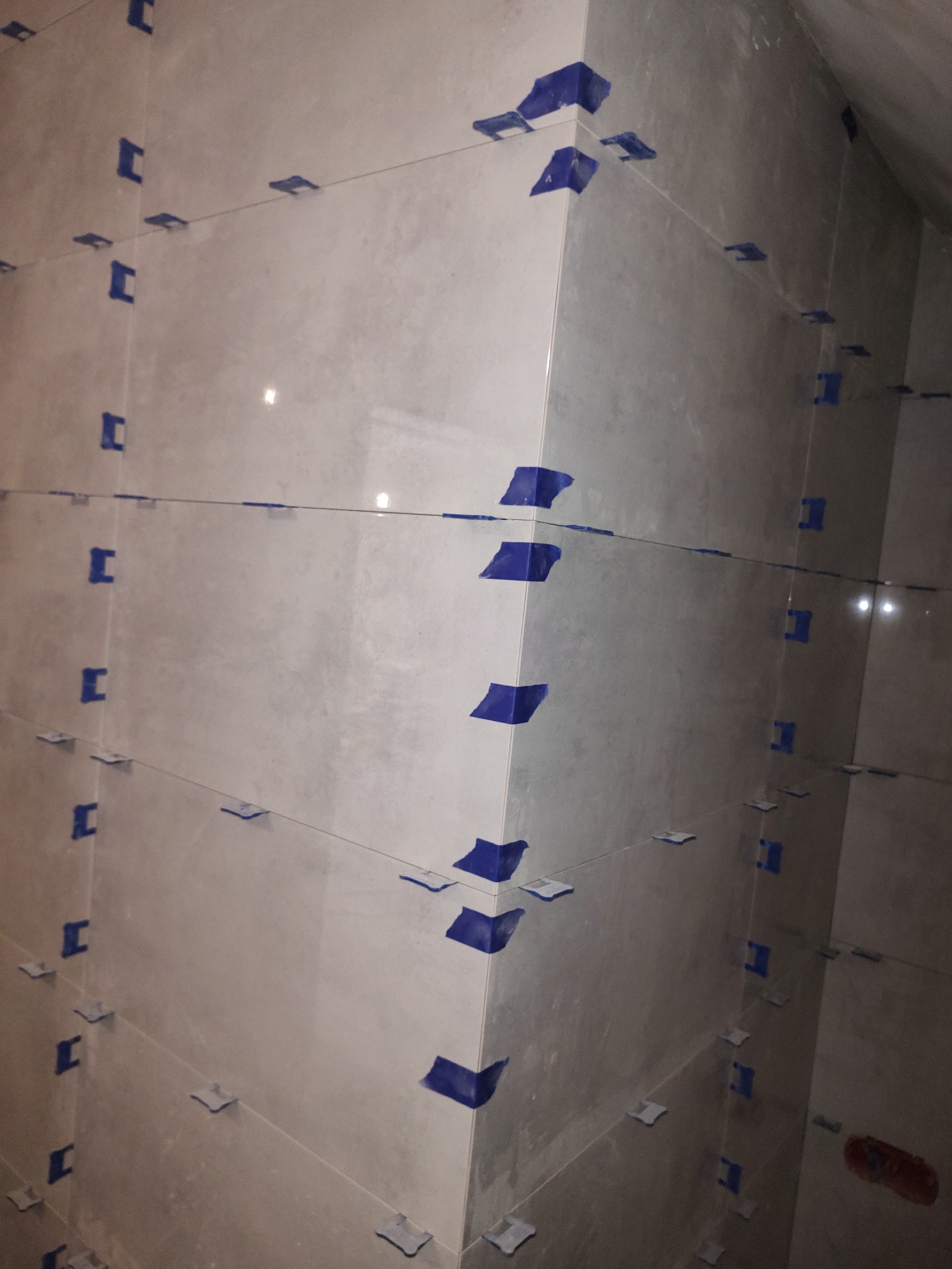 Ściana wyłożona jasnymi płytkami, z niebieskimi elementami poziomowania i sznurkiem wyznaczającym linię, ukazująca etap prac glazurniczych w rogu pomieszczenia.