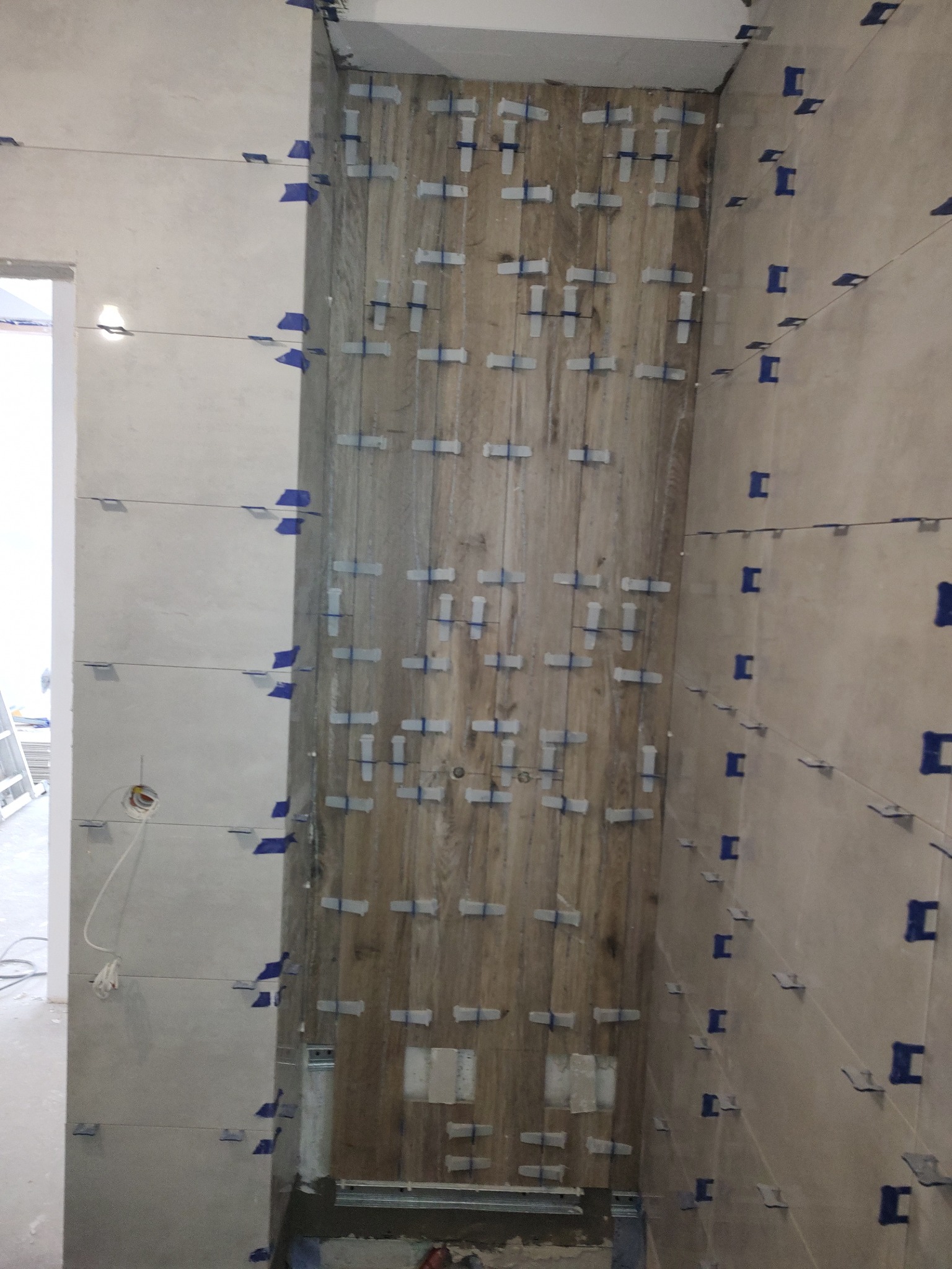 Ściana w trakcie układania płytek imitujących drewno, z użyciem krzyżyków dystansowych i poziomicy laserowej, widoczne niedokończone fragmenty i przewody elektryczne.