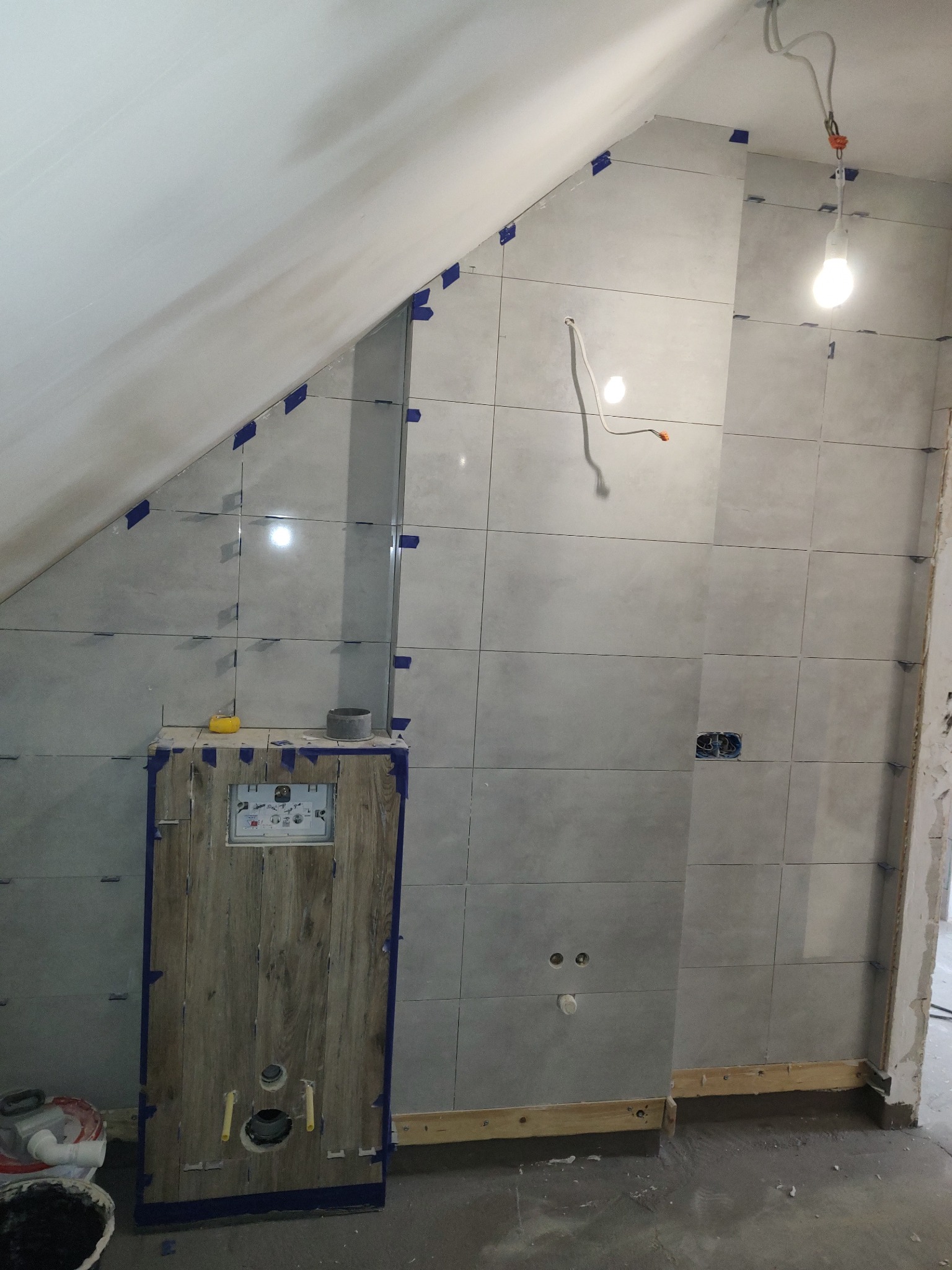 Ściana w łazience w trakcie układania szarych płytek, zabezpieczonych niebieską taśmą. Widoczna zabudowa stelaża WC z drewnopodobną okleiną i wystające kable.