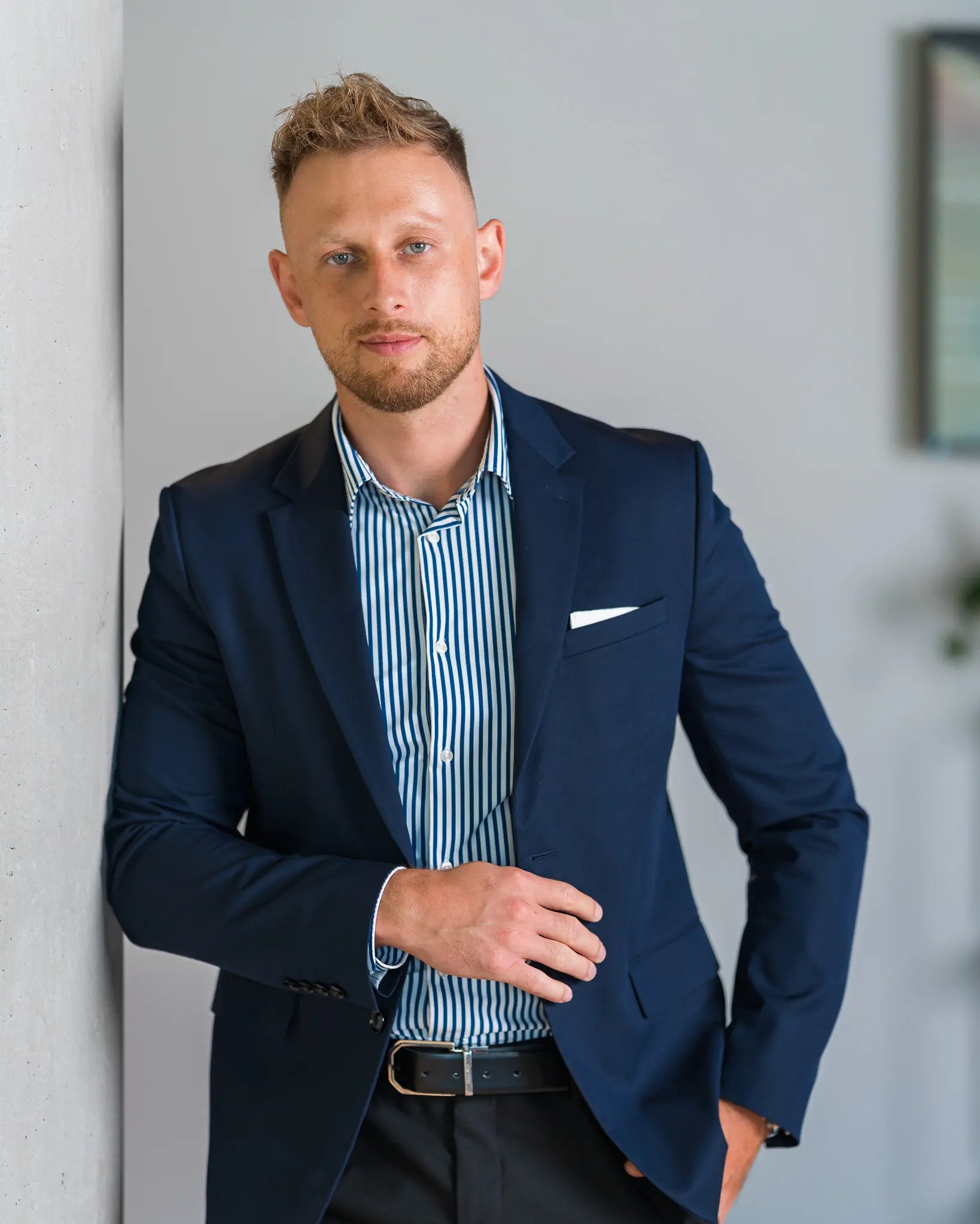 Mężczyzna w granatowym garniturze i koszuli w paski, oparty o szarą ścianę. Uwagę przykuwa jego pewny wzrok i profesjonalny wygląd.
