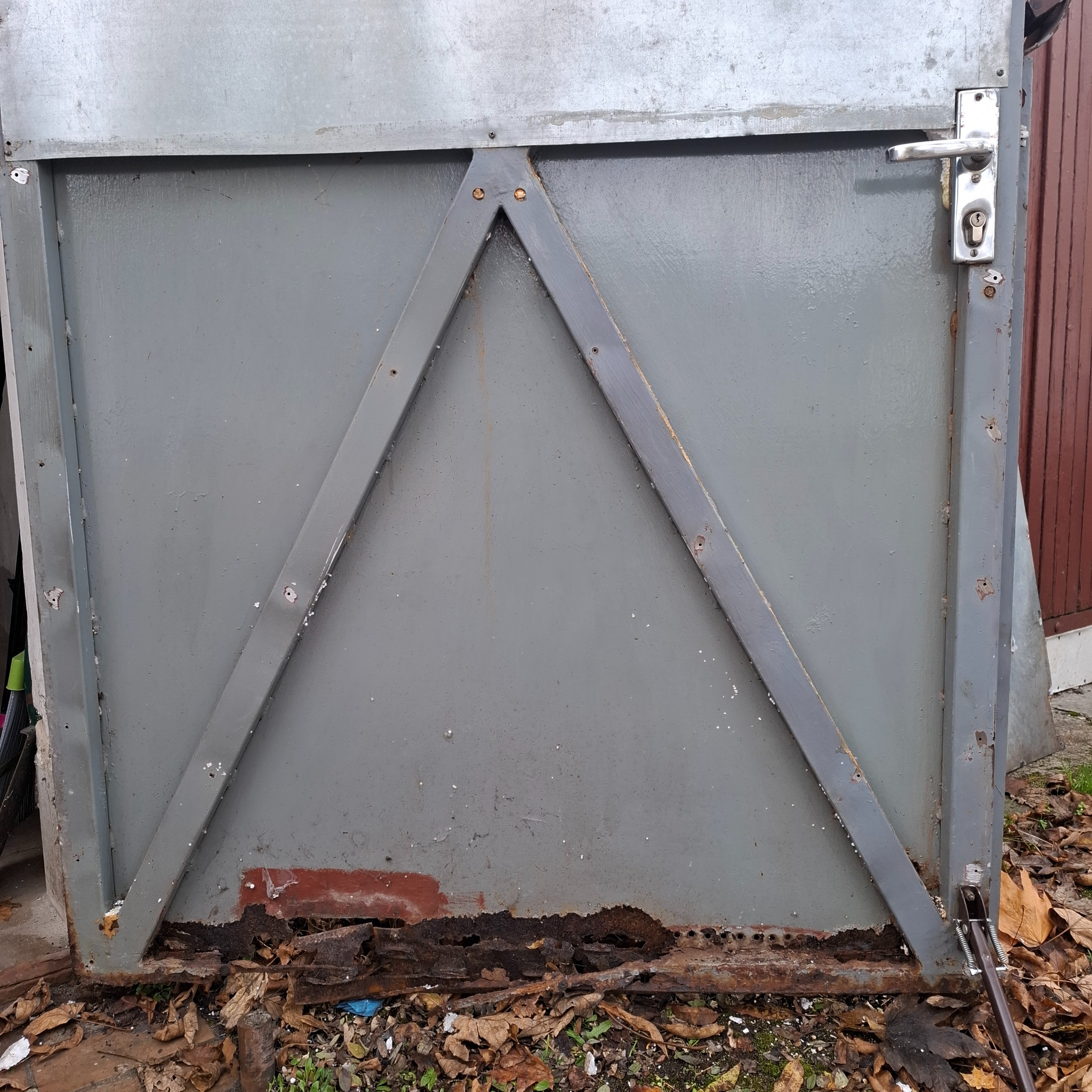 Szara, metalowa brama garażowa z widocznymi śladami korozji u dołu i wzmocnieniem w kształcie trójkąta. Widoczna klamka i zamek. Na dole leżą liście.