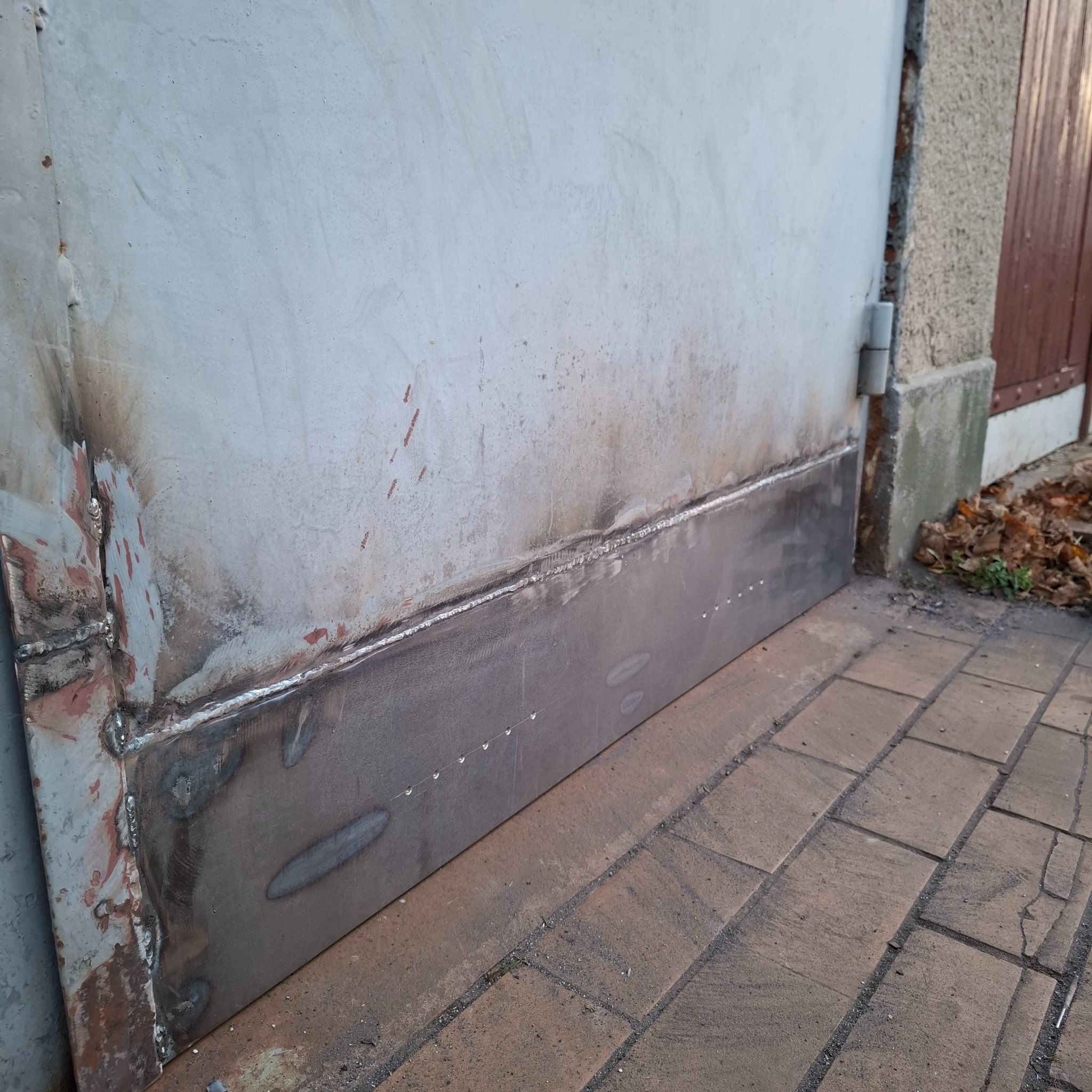 Dolna część metalowej bramy po naprawie, widoczne spawy i nowa blacha. Brama pomalowana na szaro, stoi na chodniku z kostki.