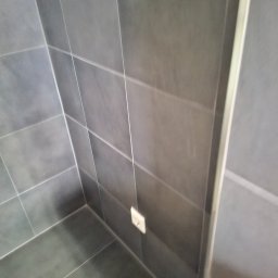 BAU-BUD SERVICE Jarosław Nawrocki - Szare, matowe płytki w kabinie prysznicowej z widocznym gniazdkiem elektrycznym i fragmentem ramy kabiny. Minimalistyczny design, nowoczesne wykończenie.