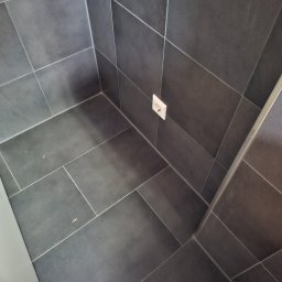 BAU-BUD SERVICE Jarosław Nawrocki - Ciemnoszare płytki podłogowe i ścienne ułożone w narożniku pomieszczenia, widoczne gniazdko elektryczne i fragment białej listwy przypodłogowej.