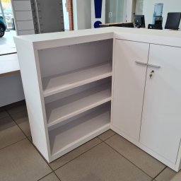 BAU-BUD SERVICE Jarosław Nawrocki - Biały, narożny regał z półkami i zamykaną szafką, widoczny na tle jasnego wnętrza z elementami industrialnymi. Nowoczesny design mebla.