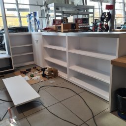 BAU-BUD SERVICE Jarosław Nawrocki - Białe regały z półkami w trakcie montażu w salonie samochodowym. Widoczne narzędzia i elementy montażowe. Jasne wnętrze z dużymi oknami i logo marki.
