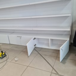 BAU-BUD SERVICE Jarosław Nawrocki - Biała zabudowa meblowa z półkami i szafkami, otwarte drzwiczki szafek dolnych, widoczne narzędzie montażowe na podłodze. Minimalistyczny design.