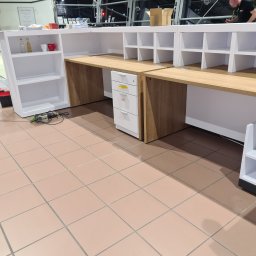 BAU-BUD SERVICE Jarosław Nawrocki - Montaż lady recepcyjnej z drewnianym blatem i białymi szafkami. Widoczne narzędzia i częściowo osoba montująca. Jasne wnętrze z przeszklonym dachem.